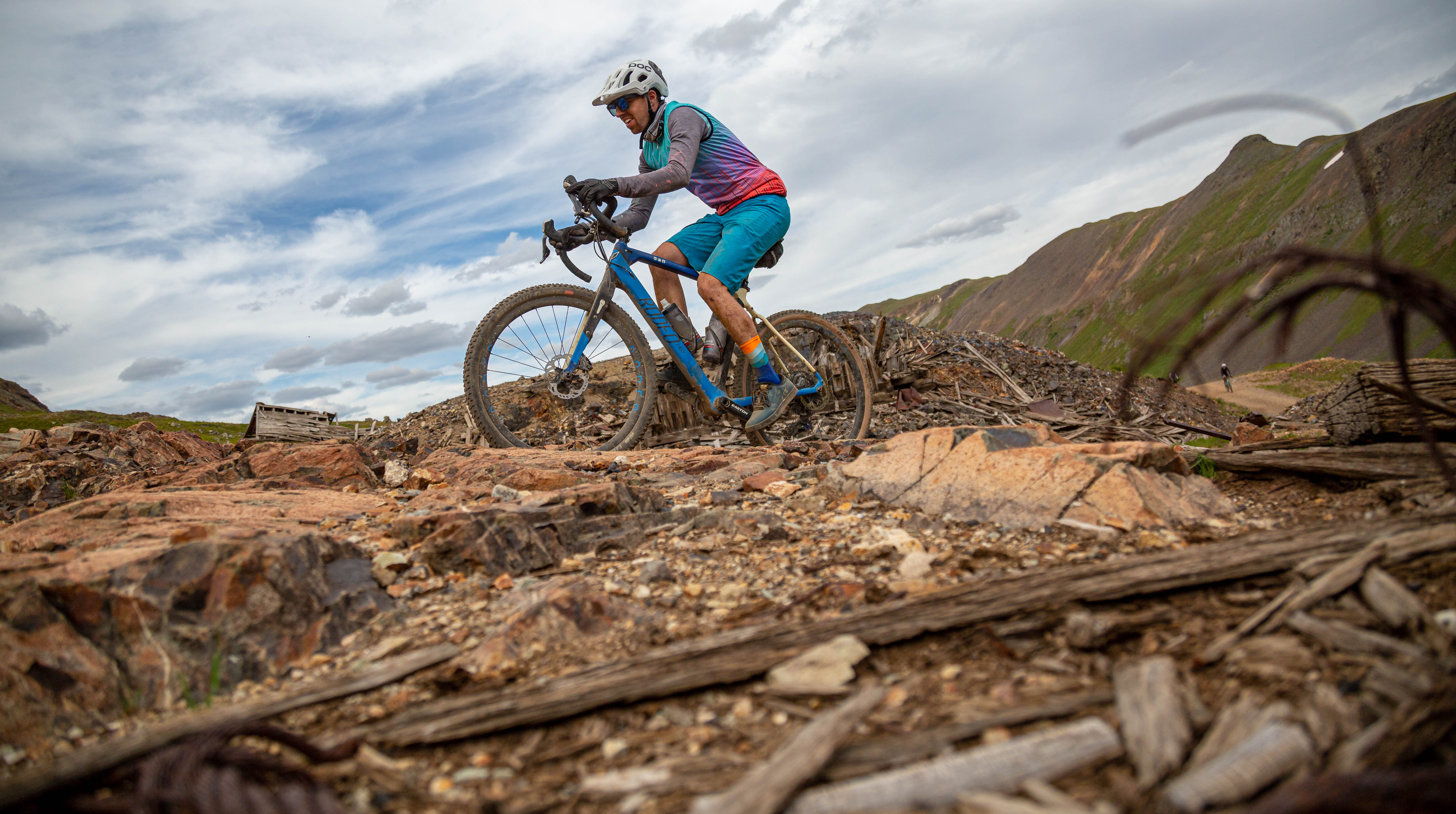 A cyclist navigating rocky terrain on a mountain bike in a scenic, mountainous landscape under a cloudy sky.