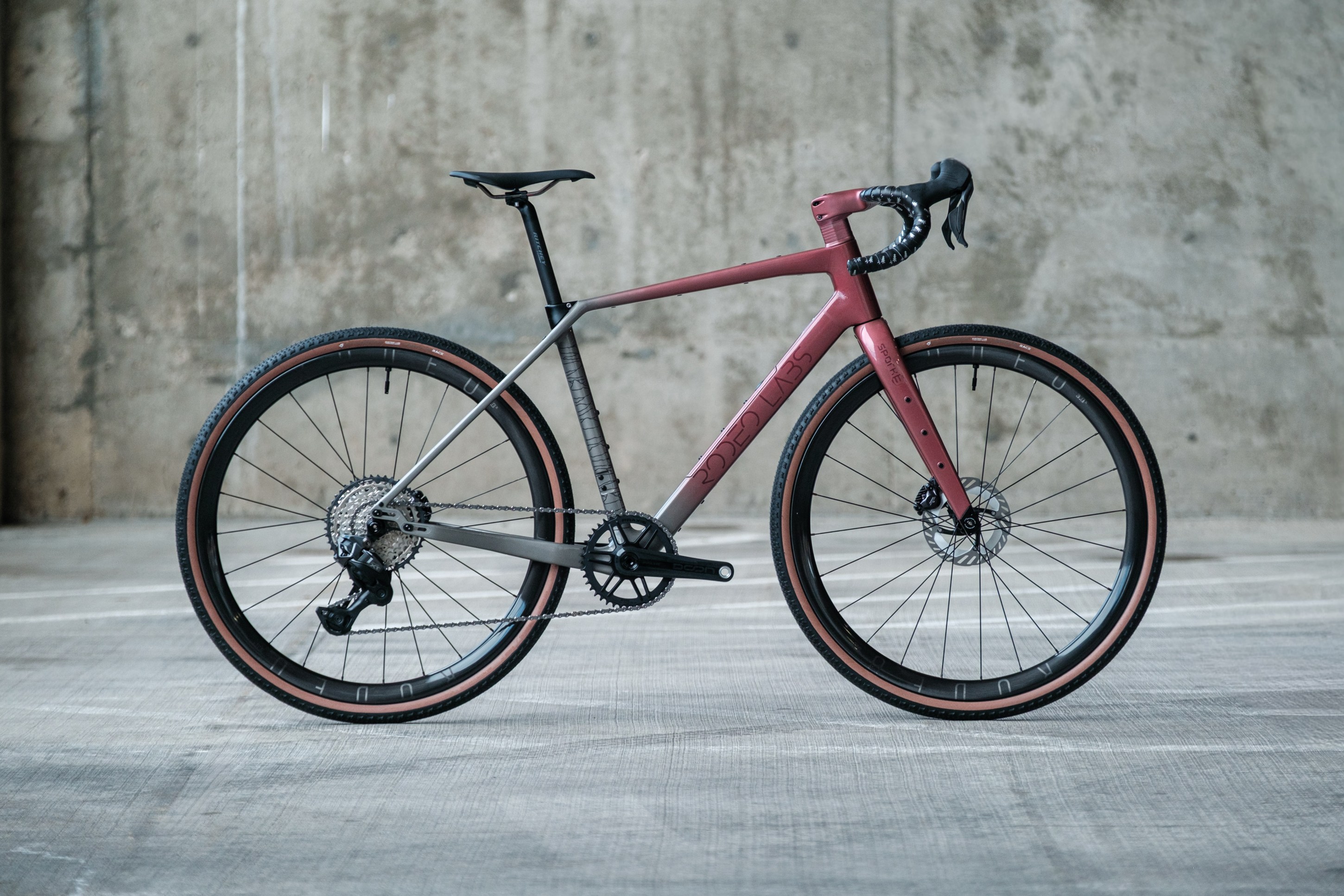 A modern gravel bike with a gray and maroon frame, featuring disc brakes and wide tires, displayed against a concrete wall.