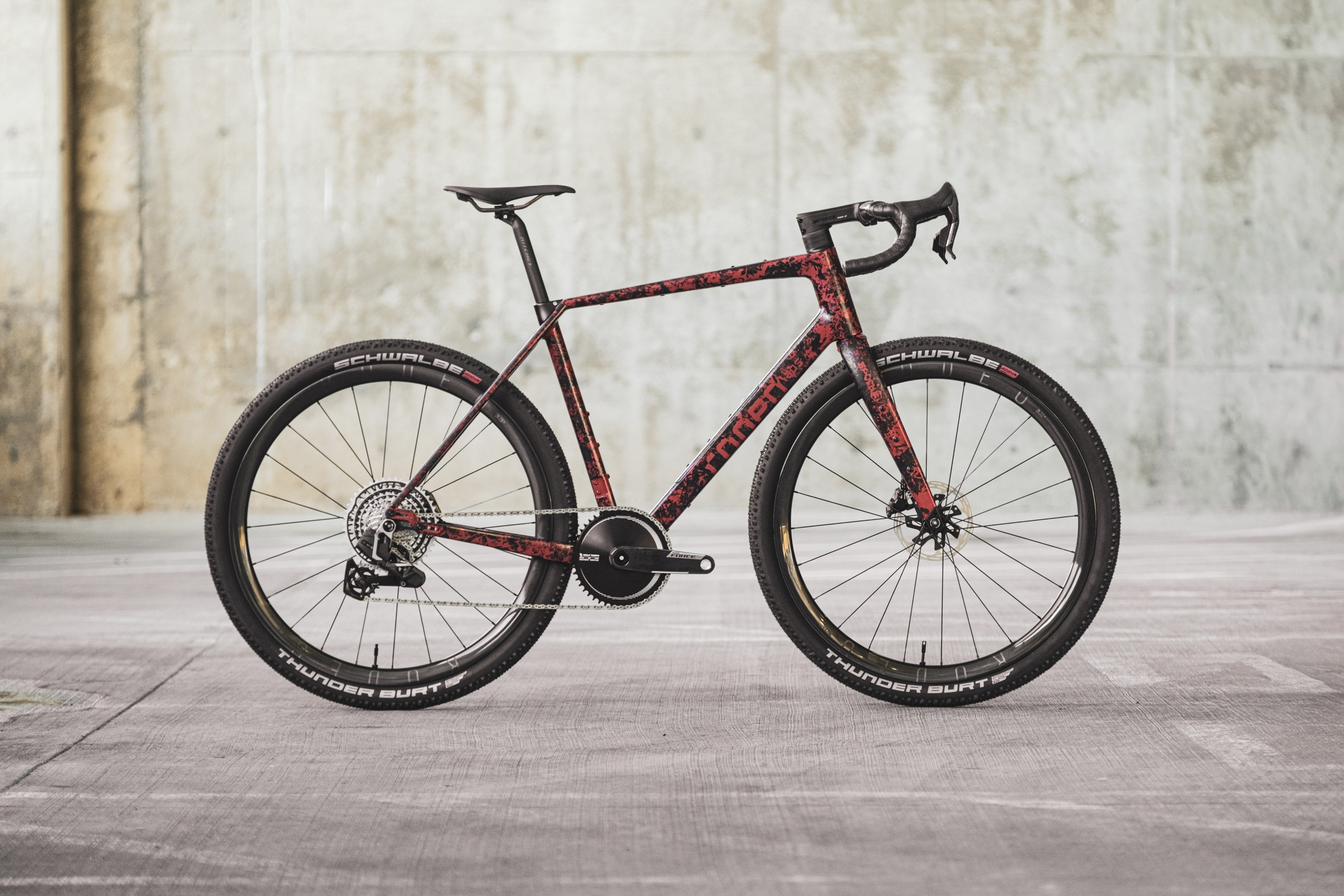 A modern bicycle with a red and black patterned frame, featuring thin tires and disc brakes, positioned against a textured concrete background.