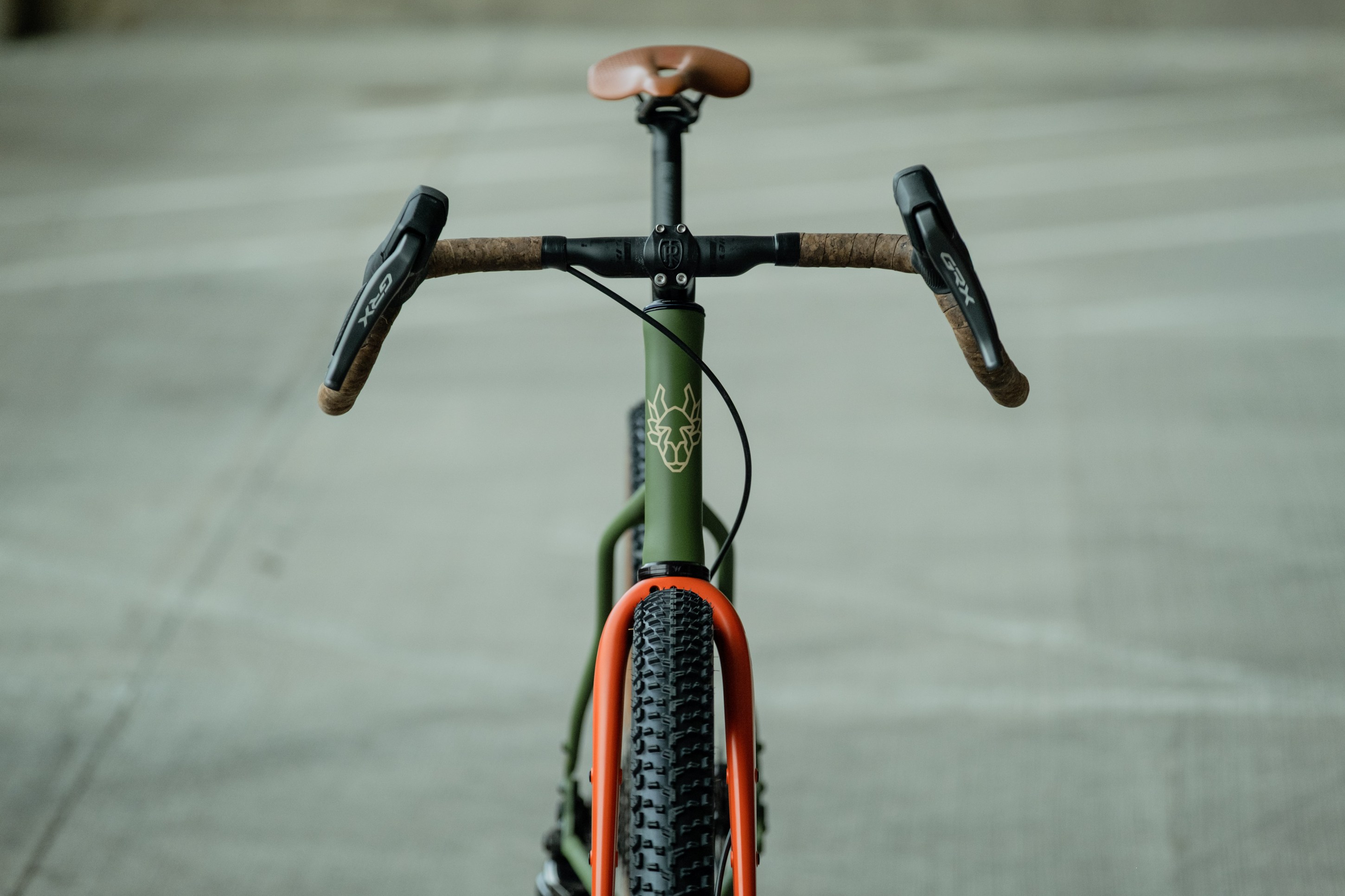 Close-up view of a bicycle's handlebars and front wheel, featuring cork grip and saddle, with an olive green frame and orange accents.