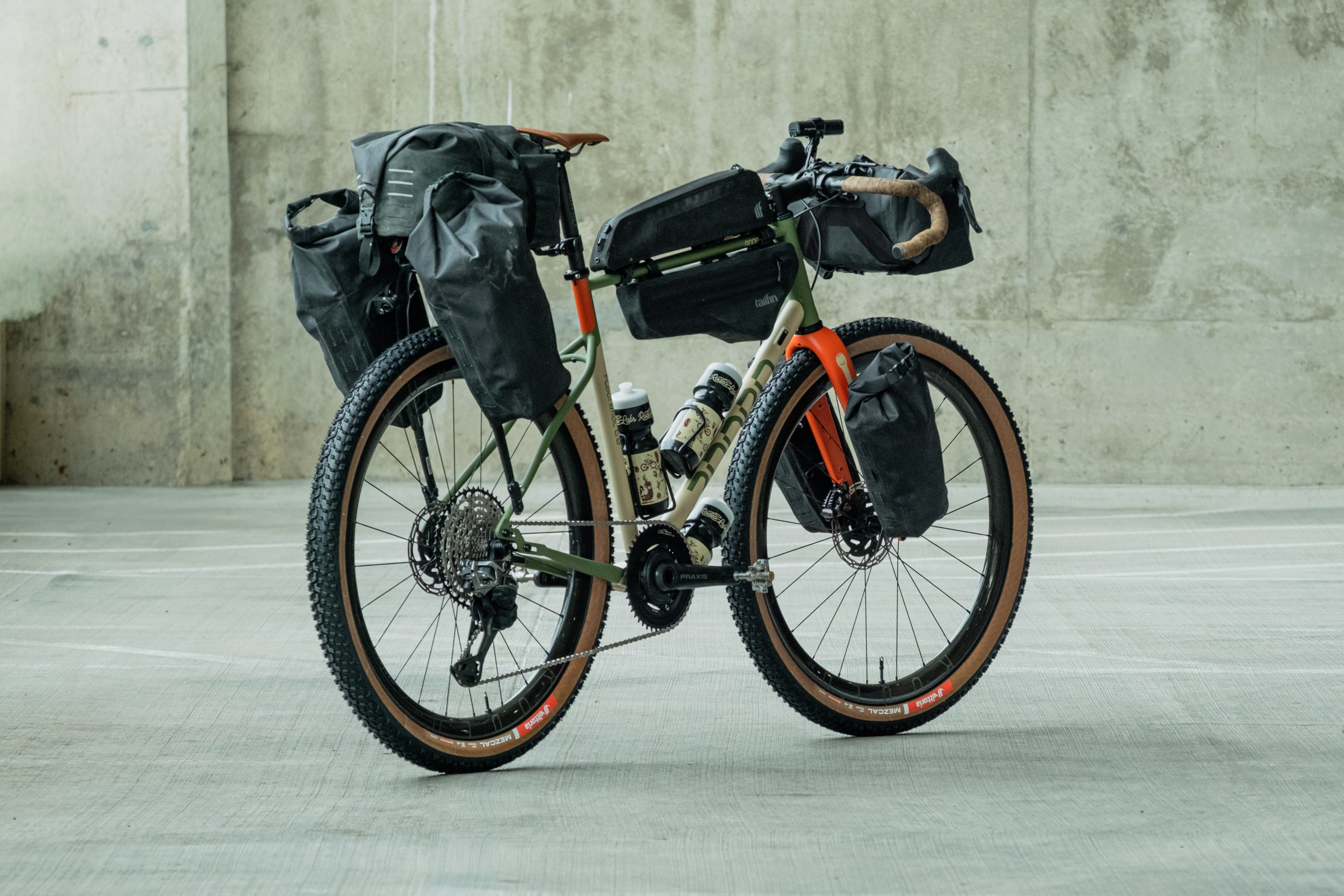 A fully loaded adventure bike with orange and green frame, featuring multiple black bags for storage, positioned on a concrete surface.
