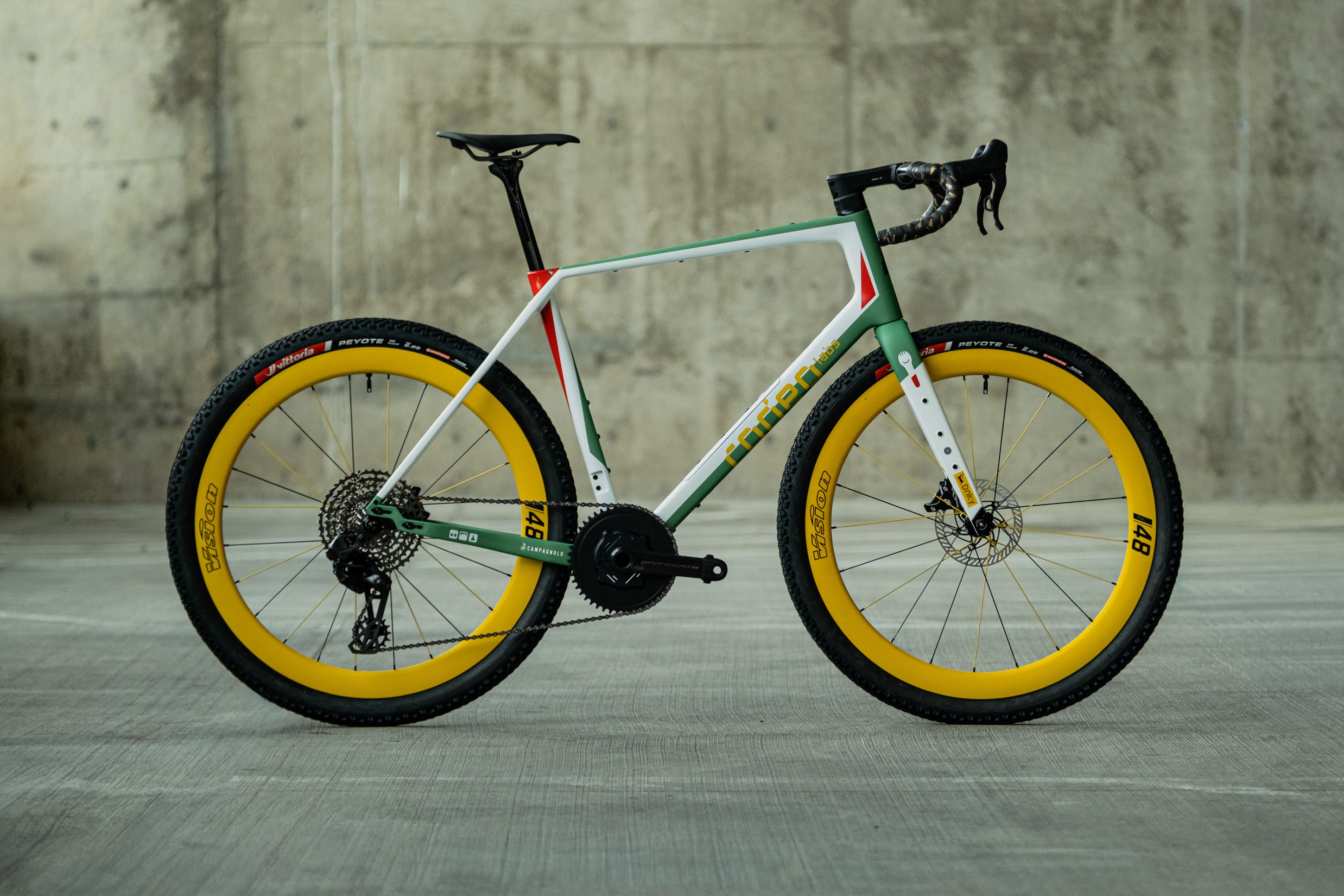 A stylish bicycle with a white, green, and red frame, featuring yellow tires and a modern design, displayed against a concrete backdrop.