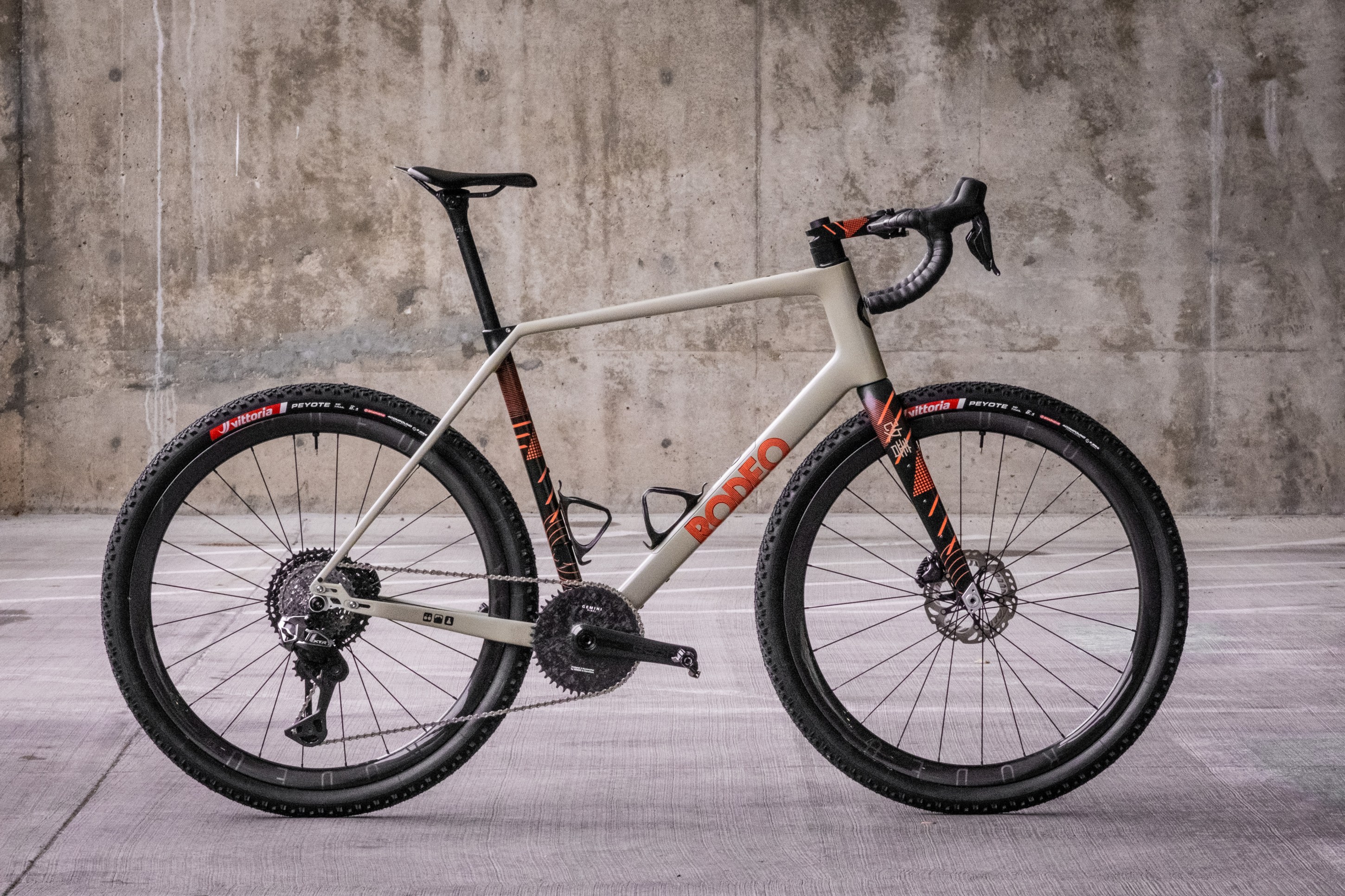 A modern gravel bike featuring a light-colored frame with orange accents, disc brakes, and rugged tires, positioned against a textured concrete wall.