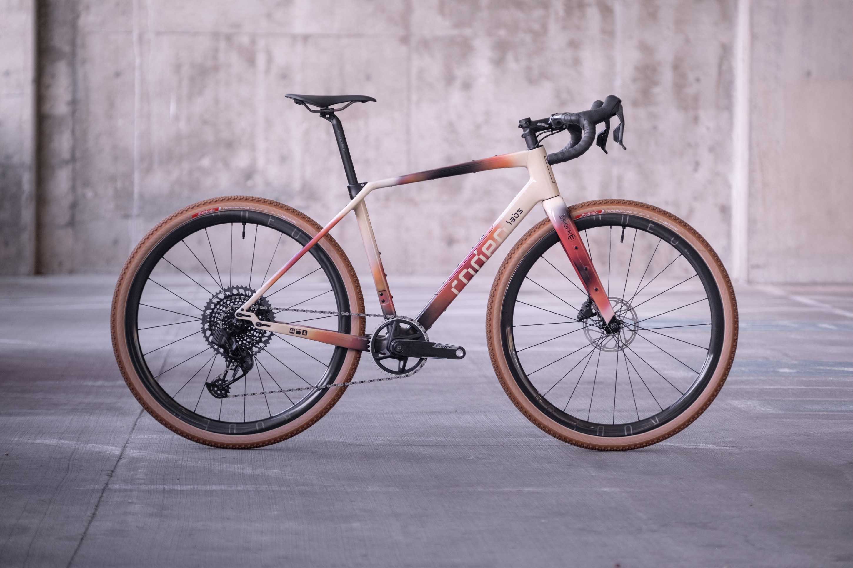 A stylish gravel bike with a gradient color frame, featuring black handlebars and tan tires, positioned in an industrial setting.