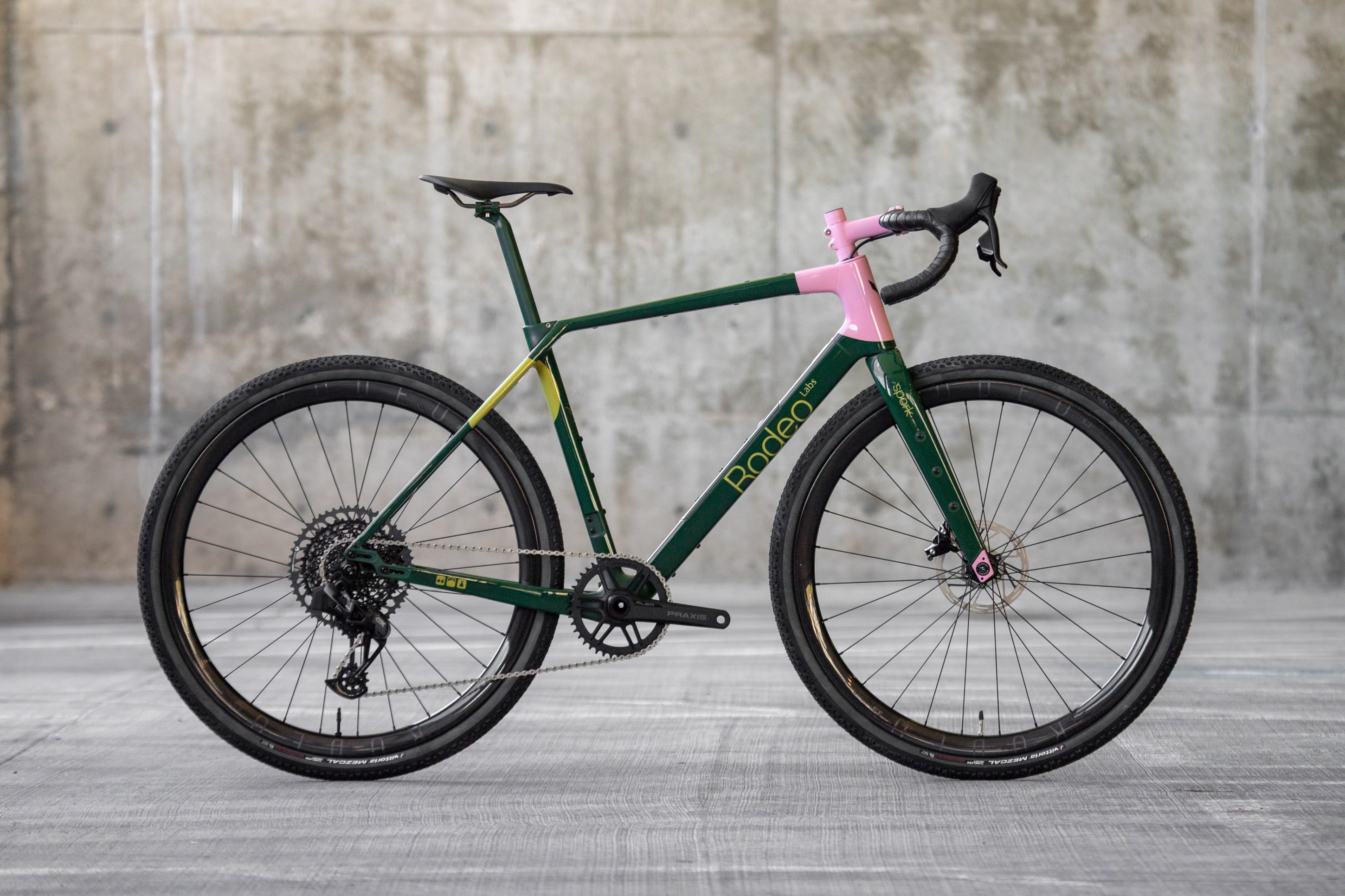 A green and pink gravel bike positioned against a concrete wall, showcasing its sleek design and high-performance features.