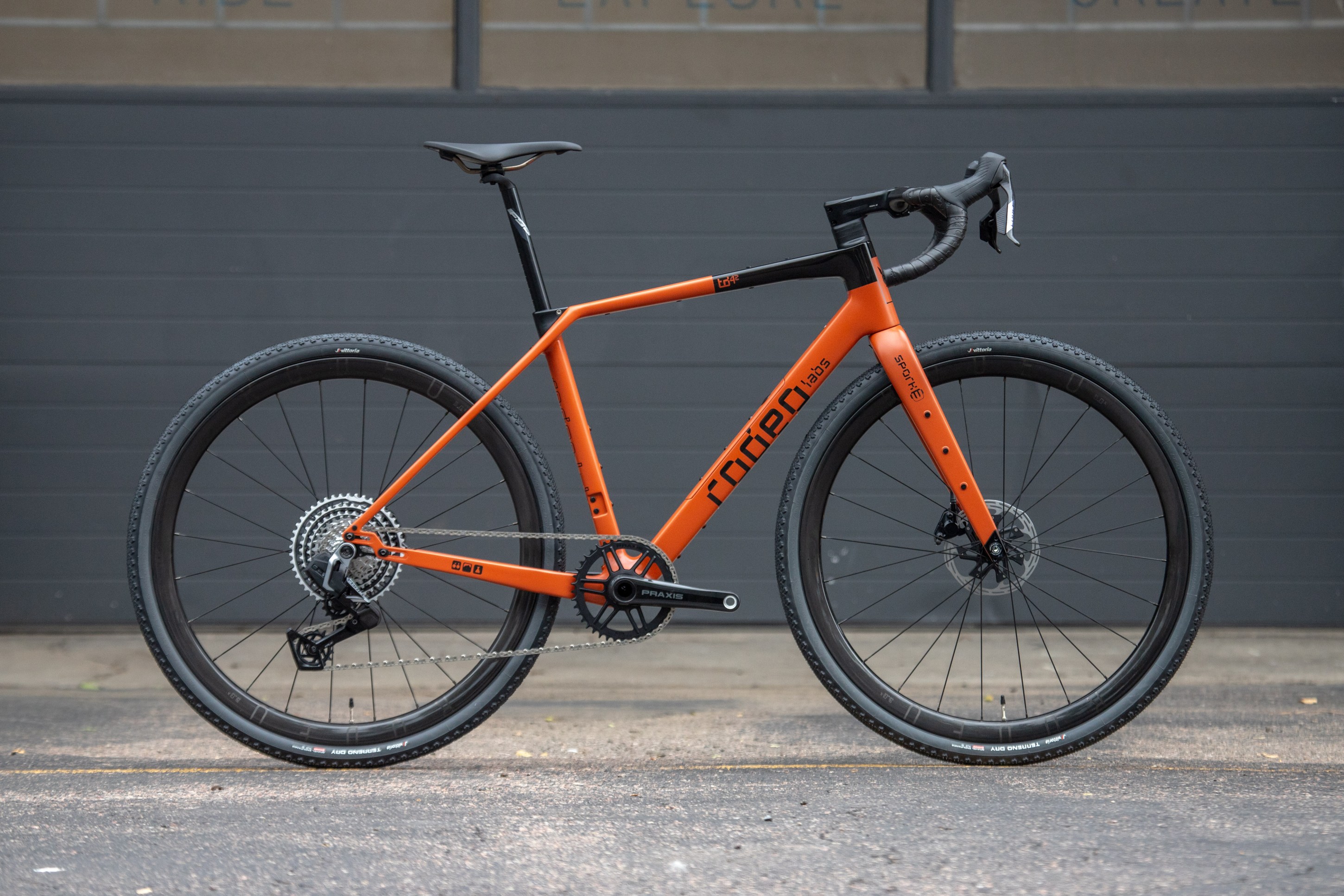 A close-up side view of an orange gravel bike showcased against a gray backdrop, featuring a sleek frame, drop handlebars, and aerodynamic design.