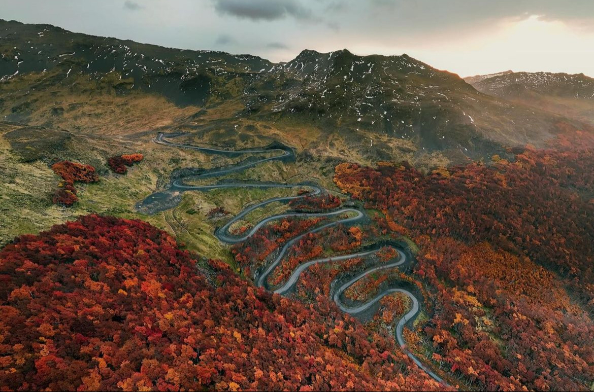Aerial view of a winding road through vibrant autumn foliage, surrounded by mountains and cloudy skies.