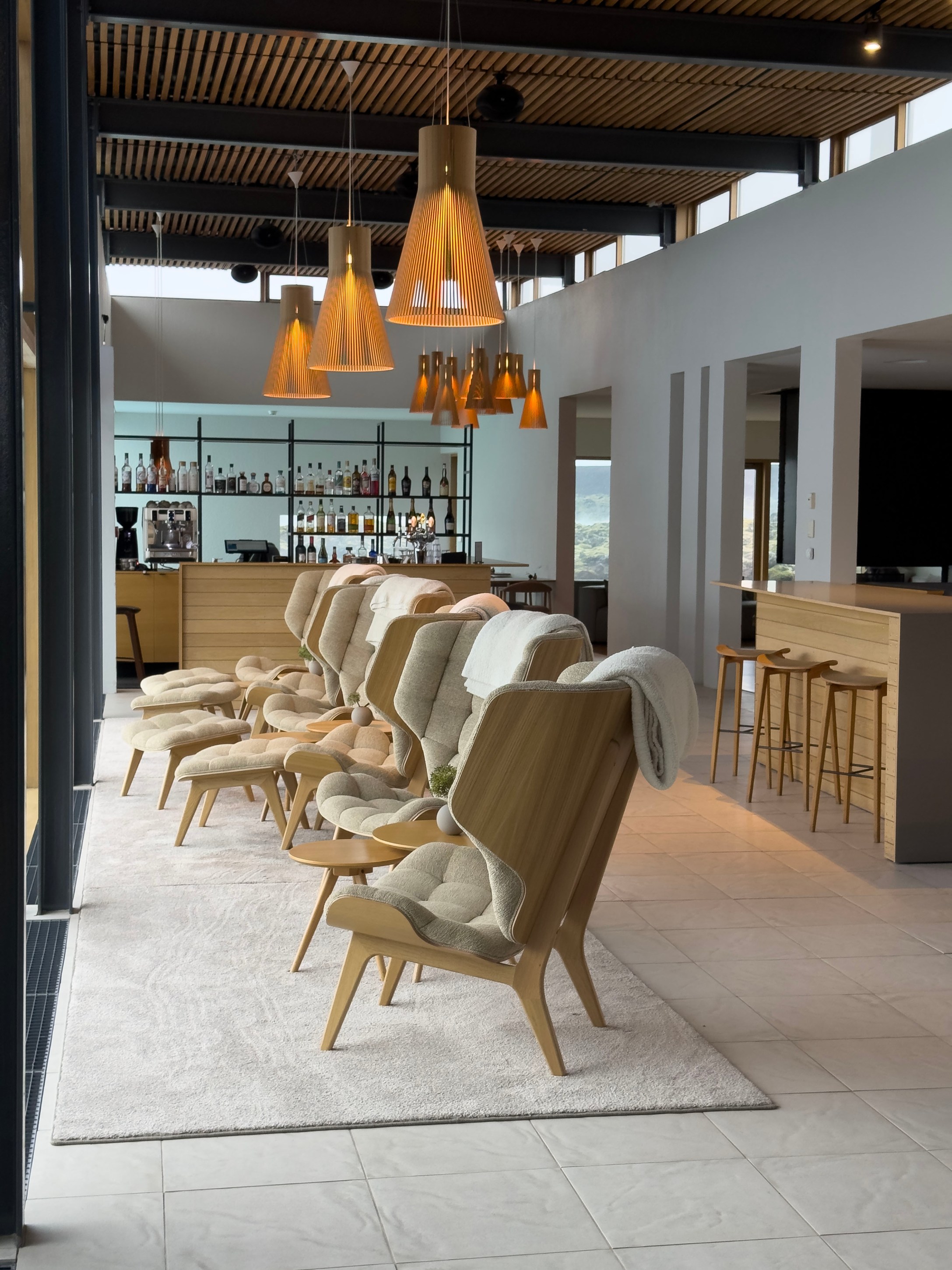 A modern lounge area featuring multiple stylish chairs with wooden frames and soft cushions, accompanied by small side tables. The space is illuminated by decorative pendant lights, and a bar area is visible in the background with various bottles. The flooring is a mix of tiles and soft rugs, creating a cozy atmosphere.