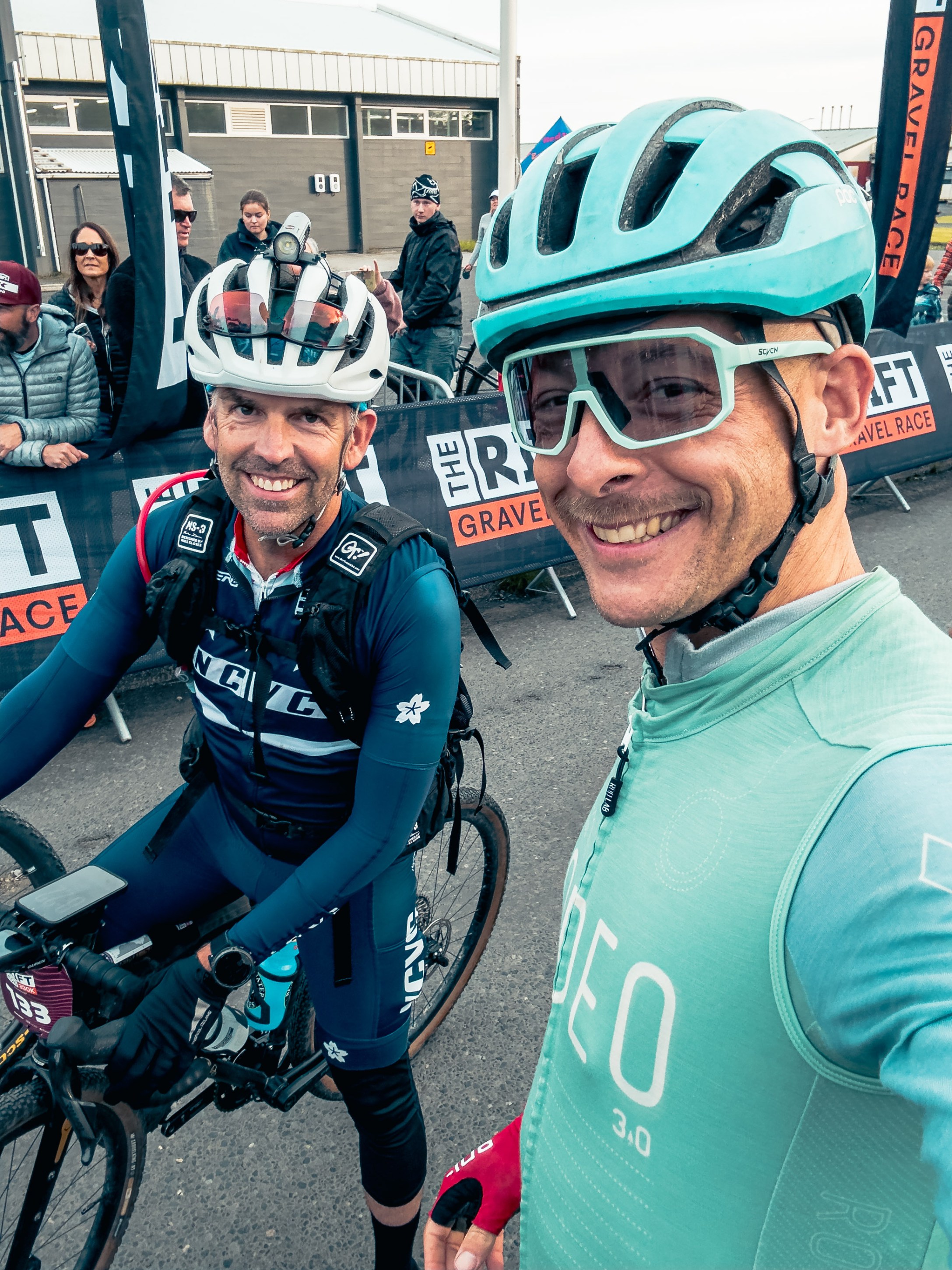 Two male cyclists posing for a selfie before a gravel race, wearing helmets and cycling gear, with a crowd in the background.