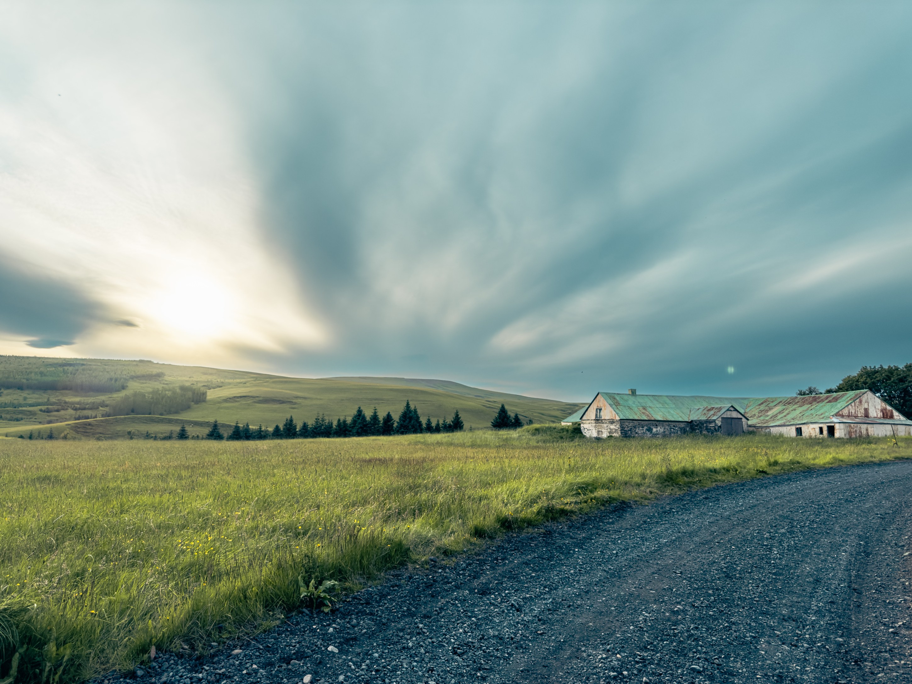 A scenic landscape featuring a gravel road leading to an old barn with a green roof, surrounded by grassy fields and rolling hills under a cloudy sky.