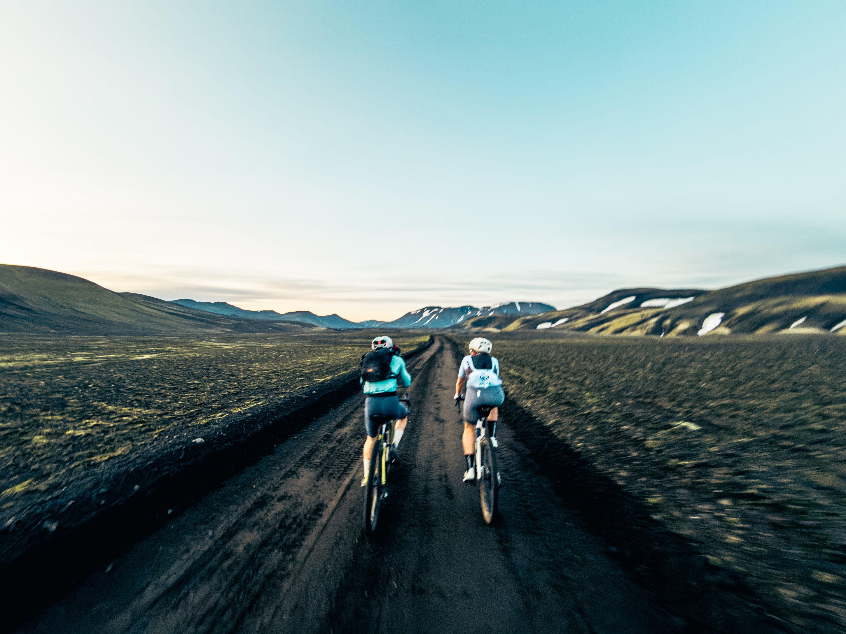 Two cyclists riding on a dirt road surrounded by mountains and a clear blue sky.