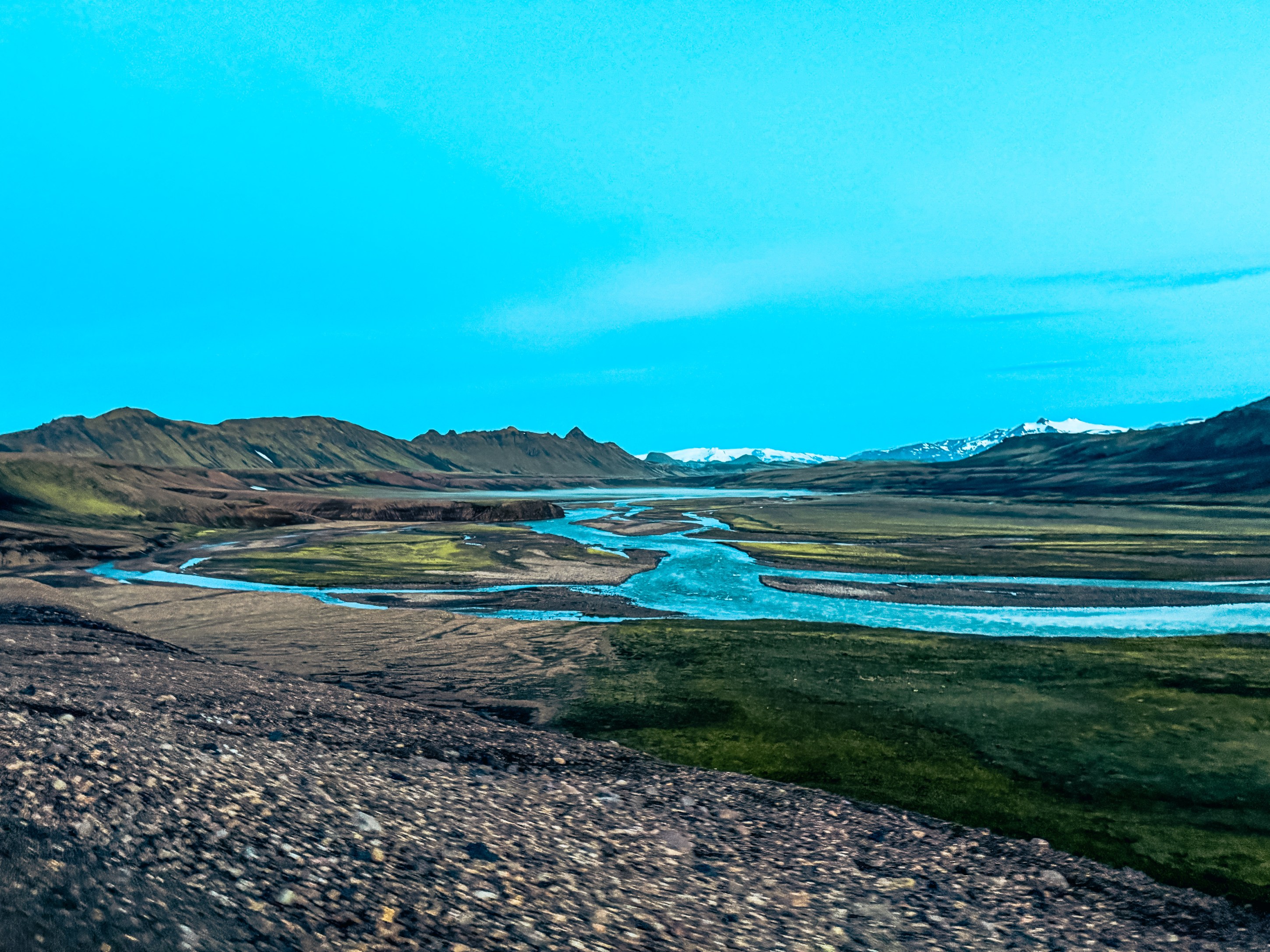 A scenic view of a winding river cutting through a lush green valley surrounded by mountains under a bright blue sky.