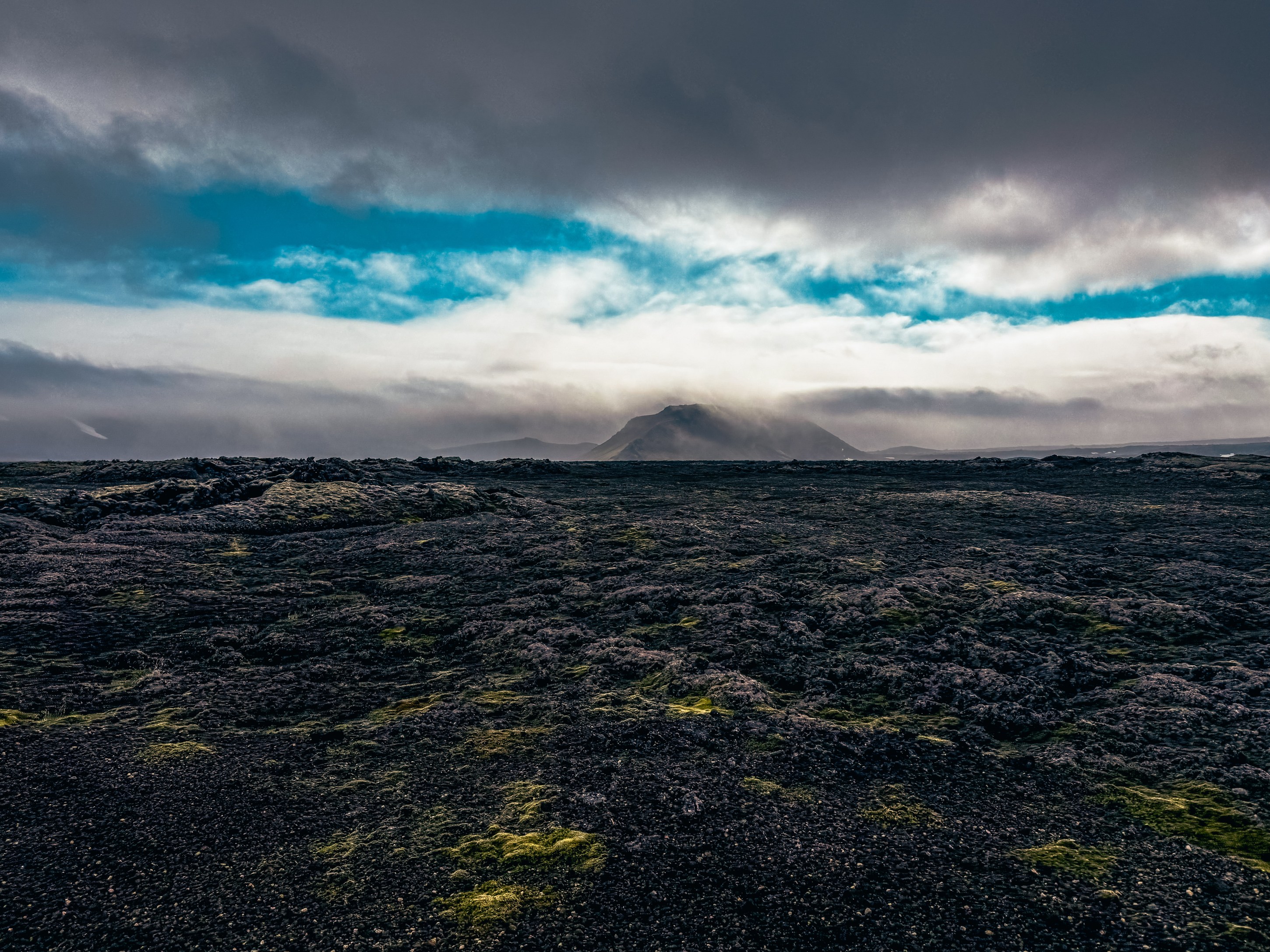A dramatic landscape featuring a black lava field with patches of green moss and a mountain in the background under a cloudy sky.