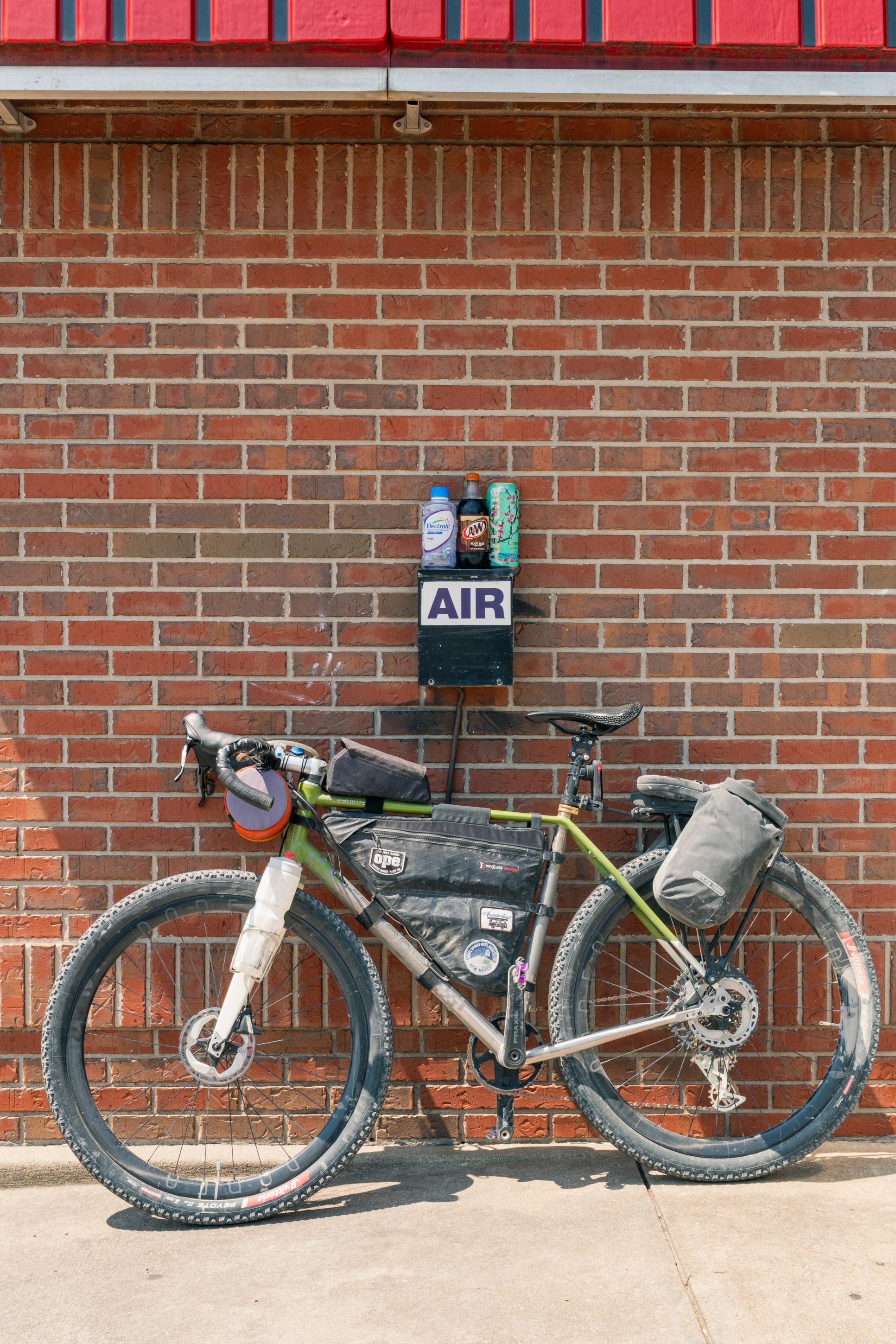 A green bicycle with various bags parked next to a brick wall, featuring a sign labeled 'AIR' above a selection of beverage bottles.
