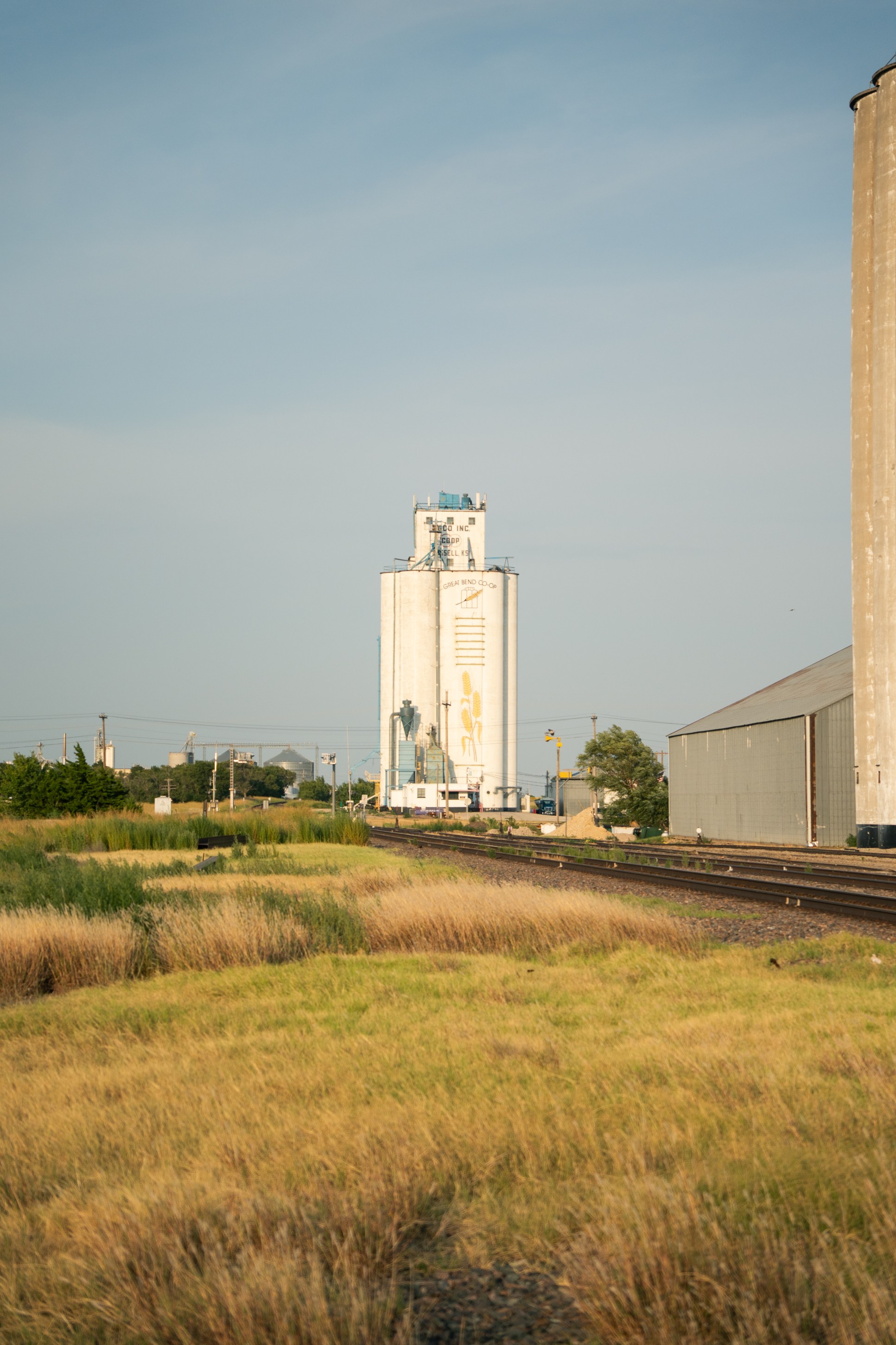 A tall grain elevator stands in the background next to railway tracks, with a grassy foreground and a clear sky.