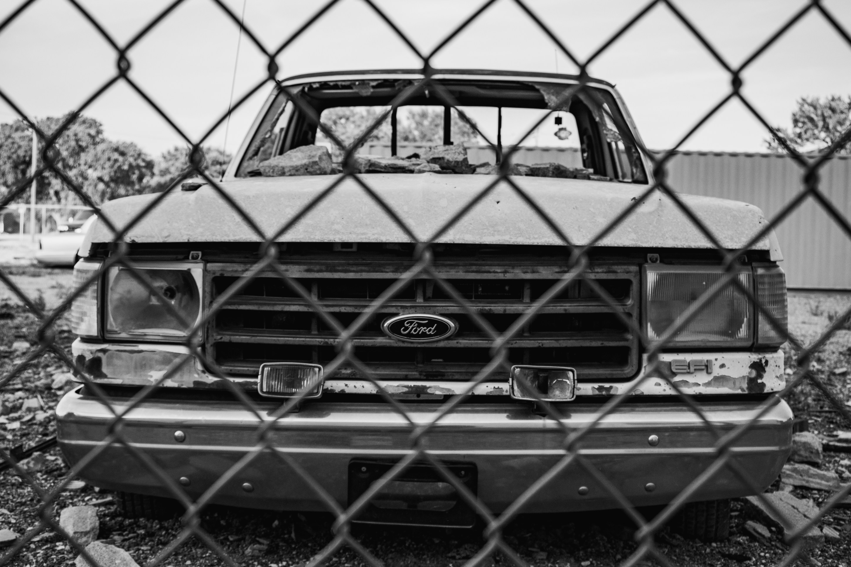 Close-up of a dilapidated Ford truck viewed through a chain-link fence, showcasing its rusty front and broken features in black and white.