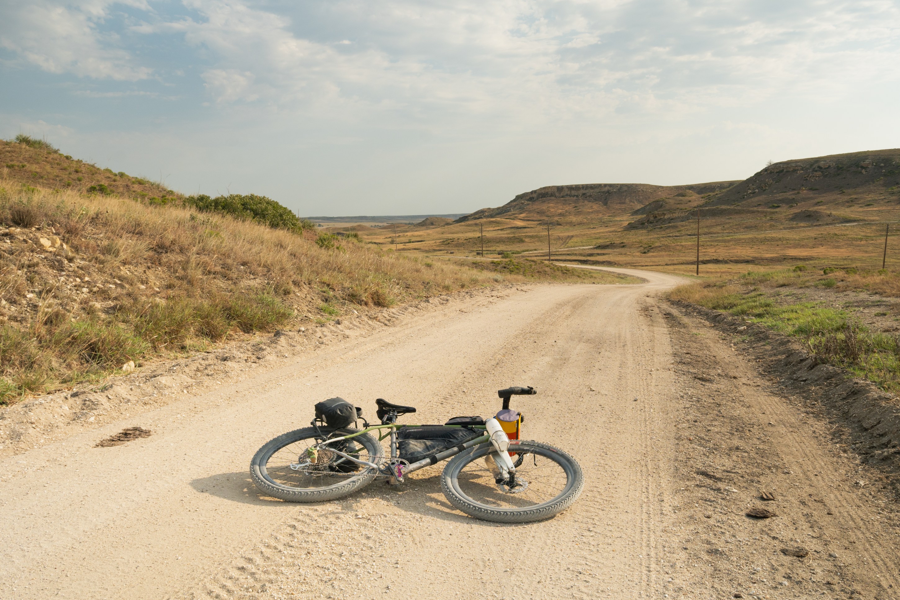 A bicycle lying on a gravel road with scenic hills in the background under a cloudy sky.