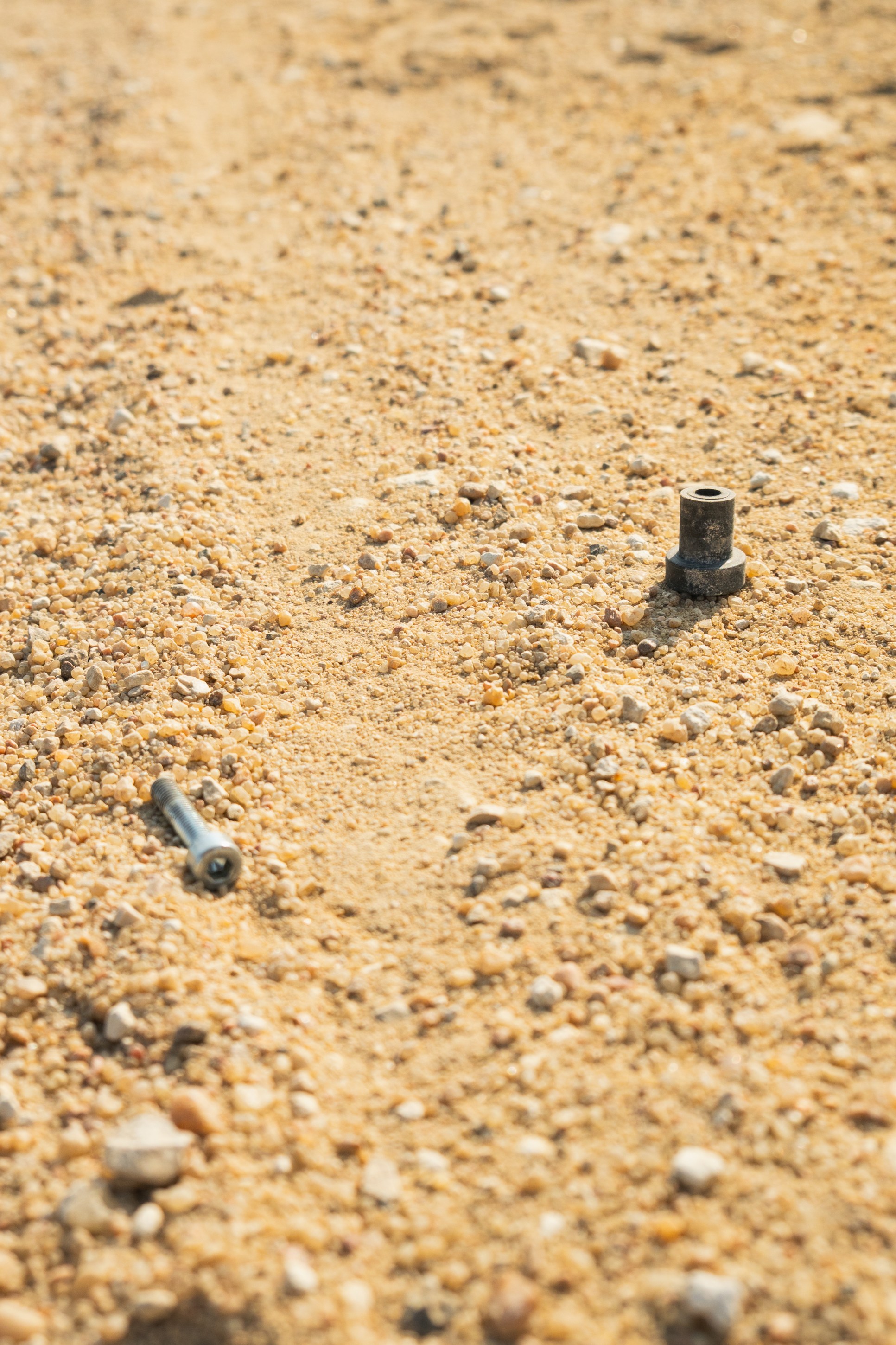 Close-up of a sandy surface with a metallic bolt and a small black connector partially buried in the sand.