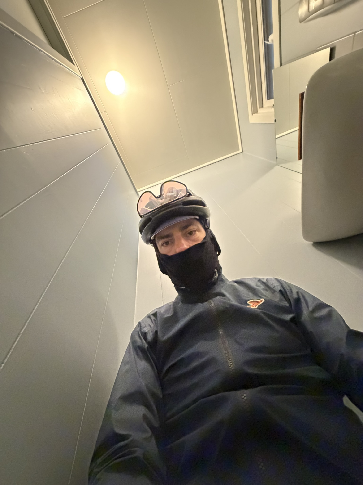 A cyclist wearing a helmet and a face mask, taking a selfie from a low angle in a room with light gray walls and a ceiling light.