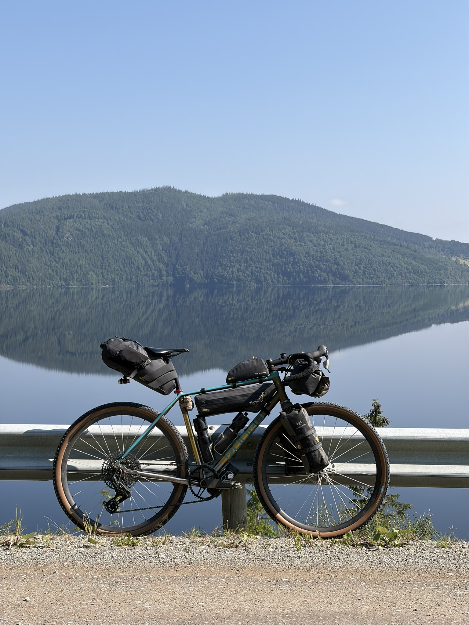 A bicycle with mounted bags parked beside a guardrail, overlooking a calm lake and lush green hills in the background under a clear blue sky.
