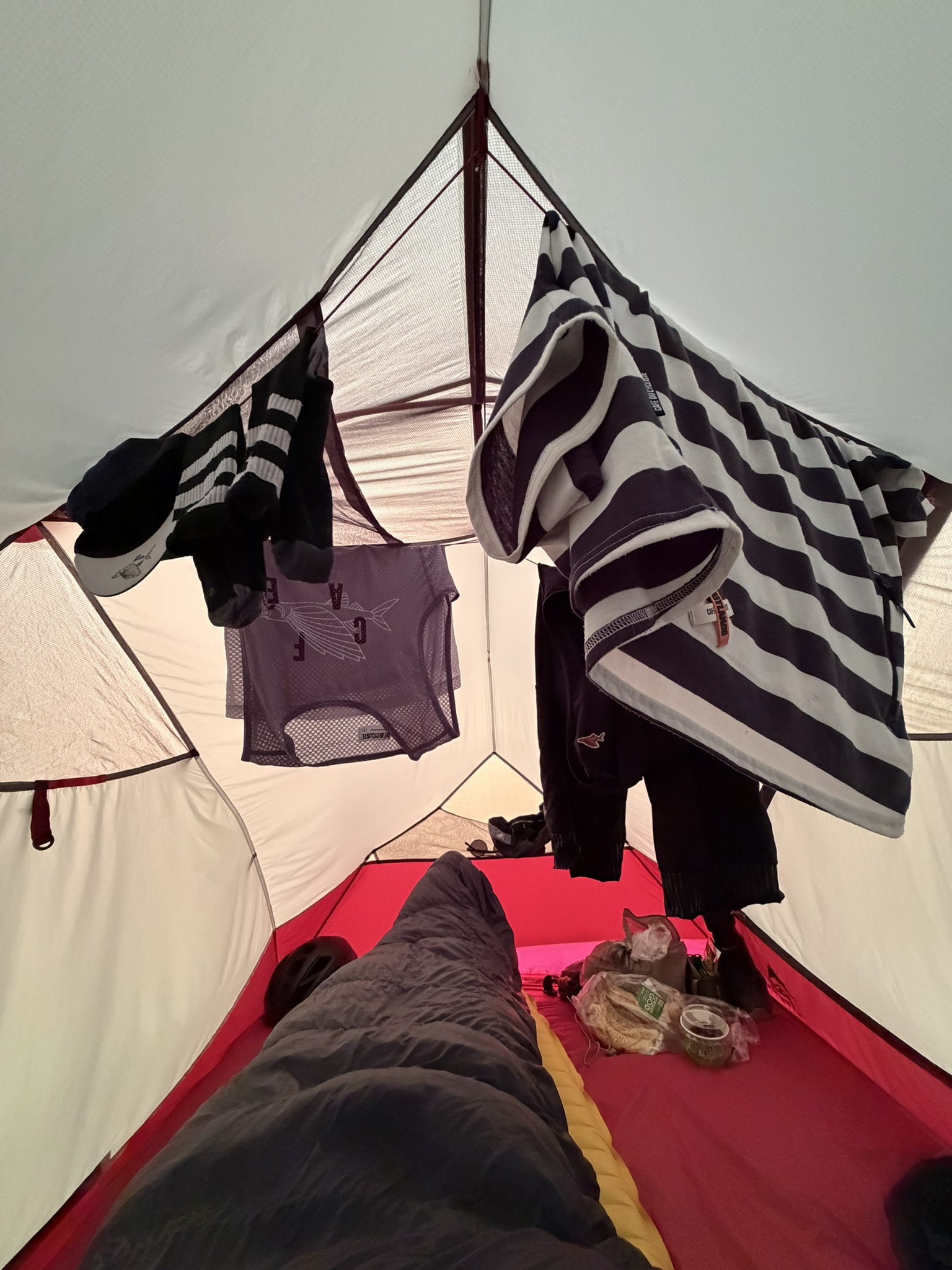 Interior of a tent with clothes hanging from the ceiling, a sleeping bag on the floor, and two cats resting on a pile of food containers.