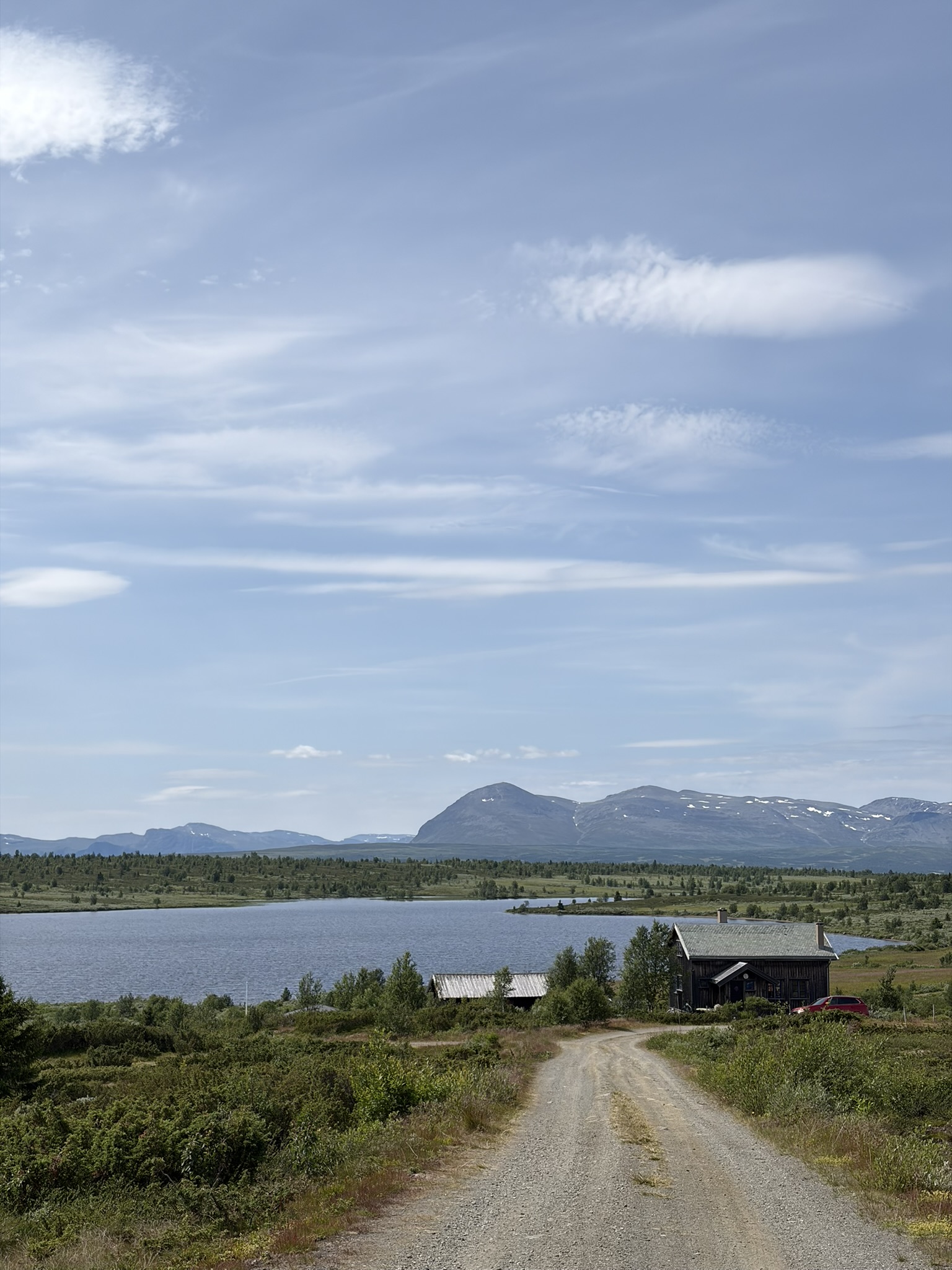 A dirt road leads to a small wooden cabin near a serene lake, surrounded by lush greenery and distant mountains under a blue sky with scattered clouds.