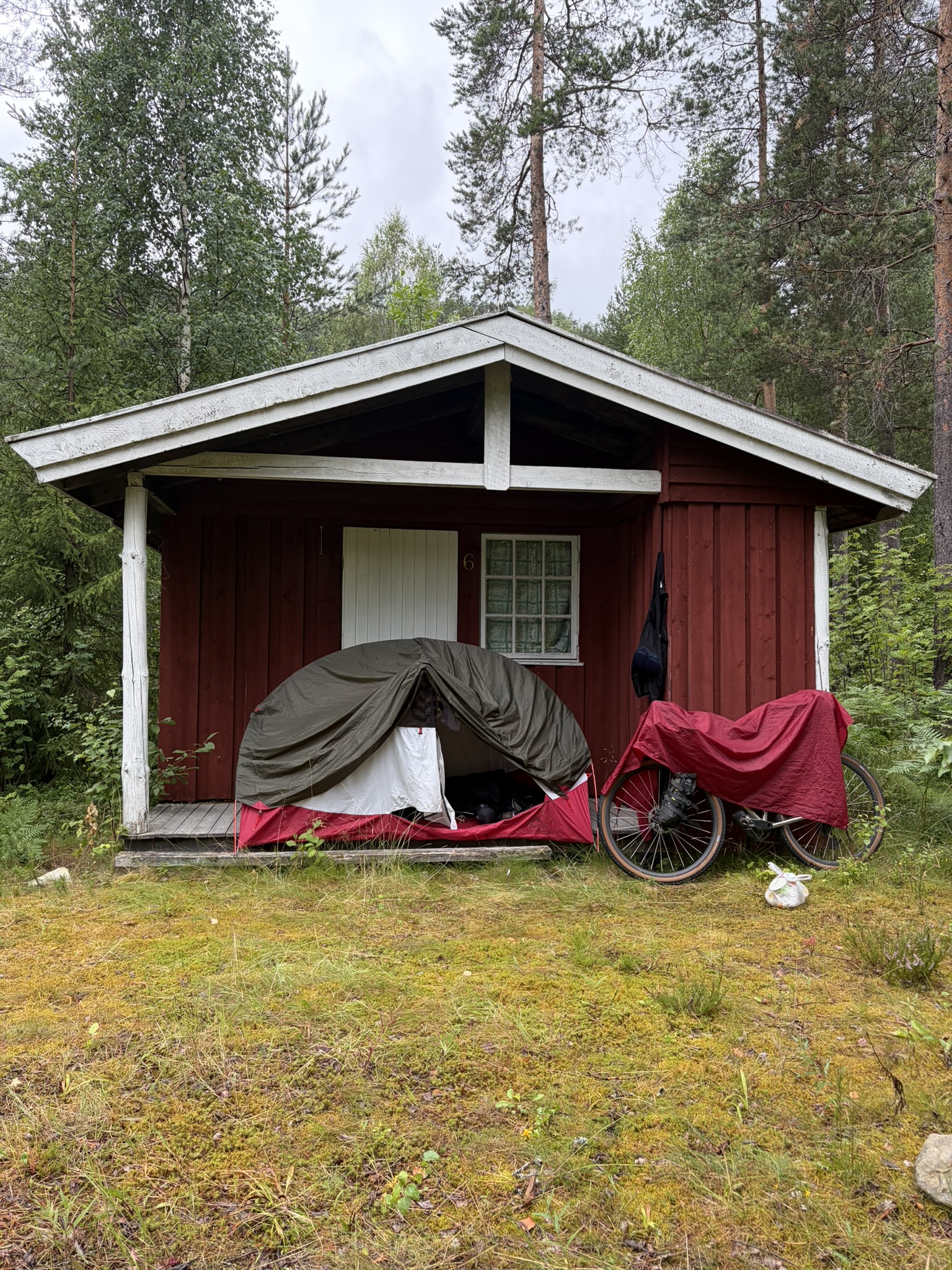 A small red wooden cabin surrounded by trees, featuring a tent set up in front and a bicycle leaning against the porch.