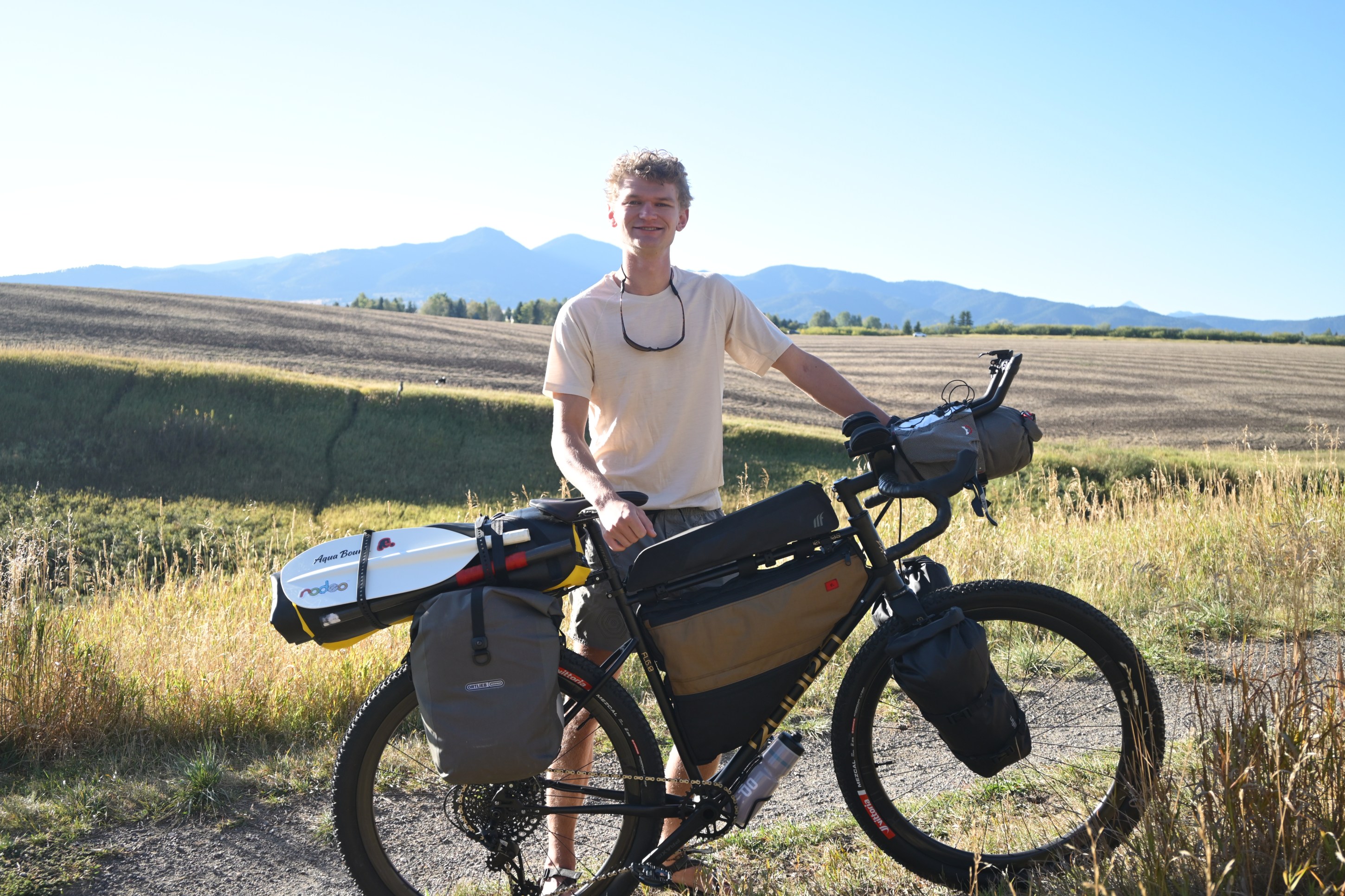 A cyclist stands beside a loaded bike in a rural setting with mountains in the background.