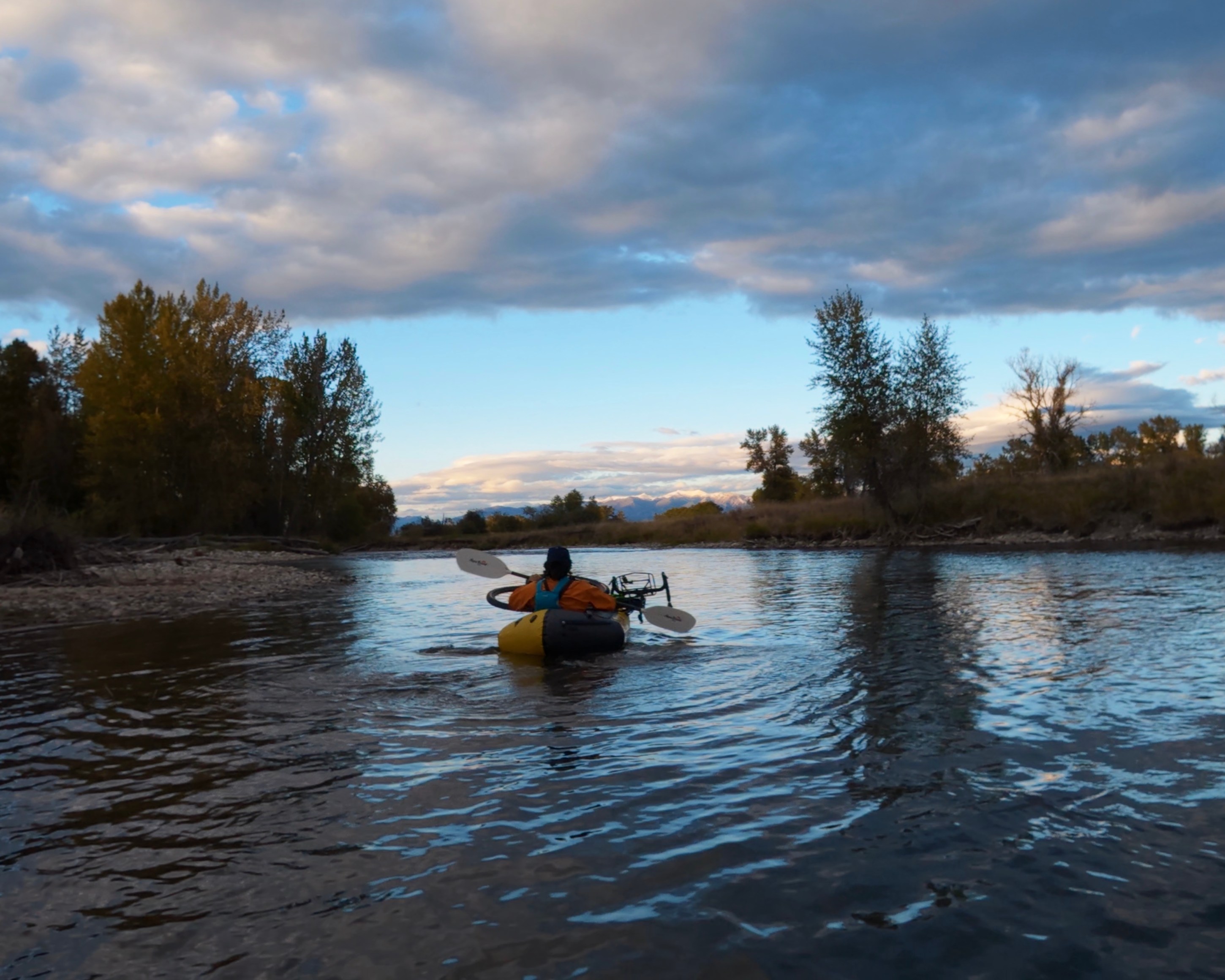 A person kayaking on a calm river surrounded by trees and clouds in the sky.