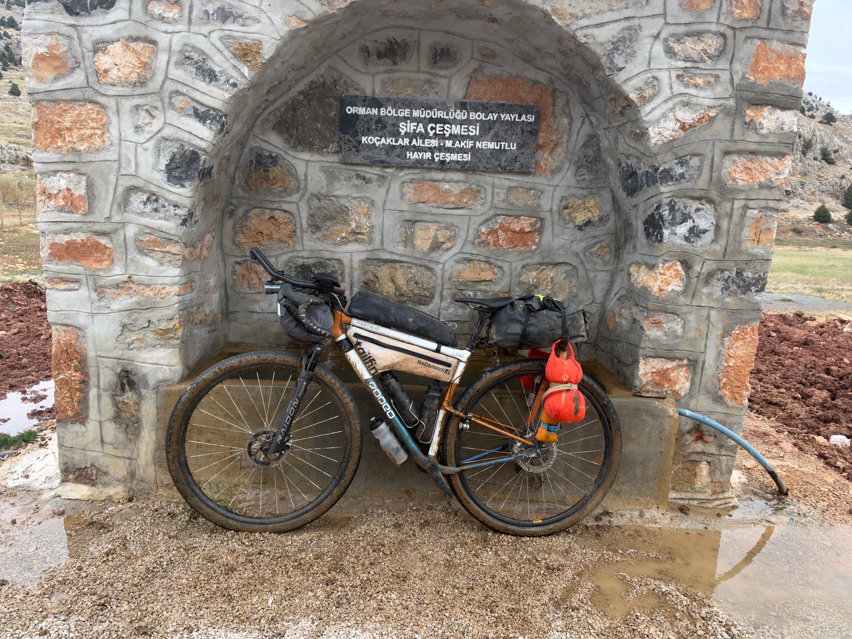 A rugged bicycle leaning against a stone structure with a sign that reads 'Sıra Çeşmesi' in Turkish, surrounded by a grassy area and muddy soil.