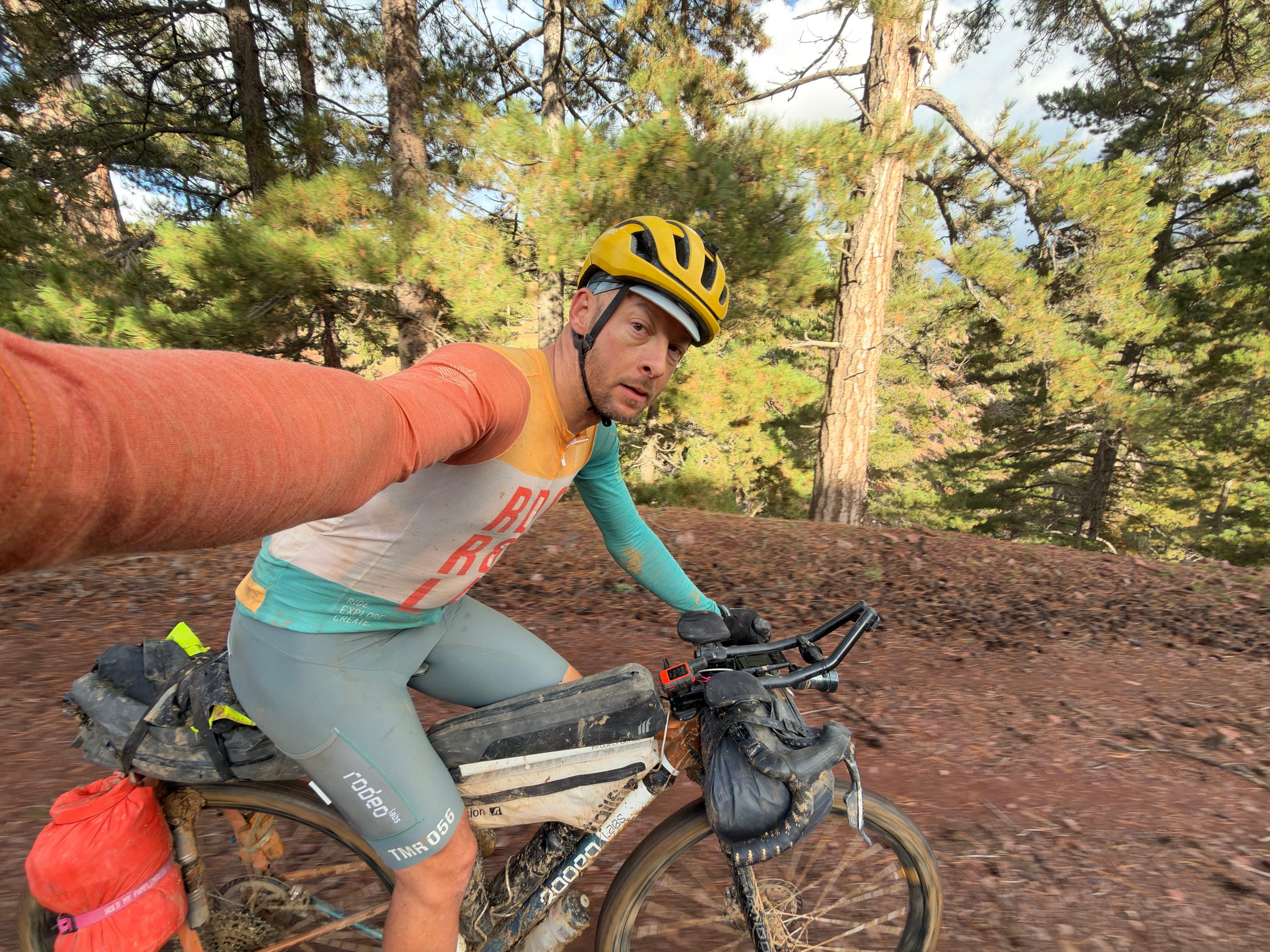 A cyclist wearing a yellow helmet and a colorful cycling jersey takes a selfie while riding a bike on a wooded trail.