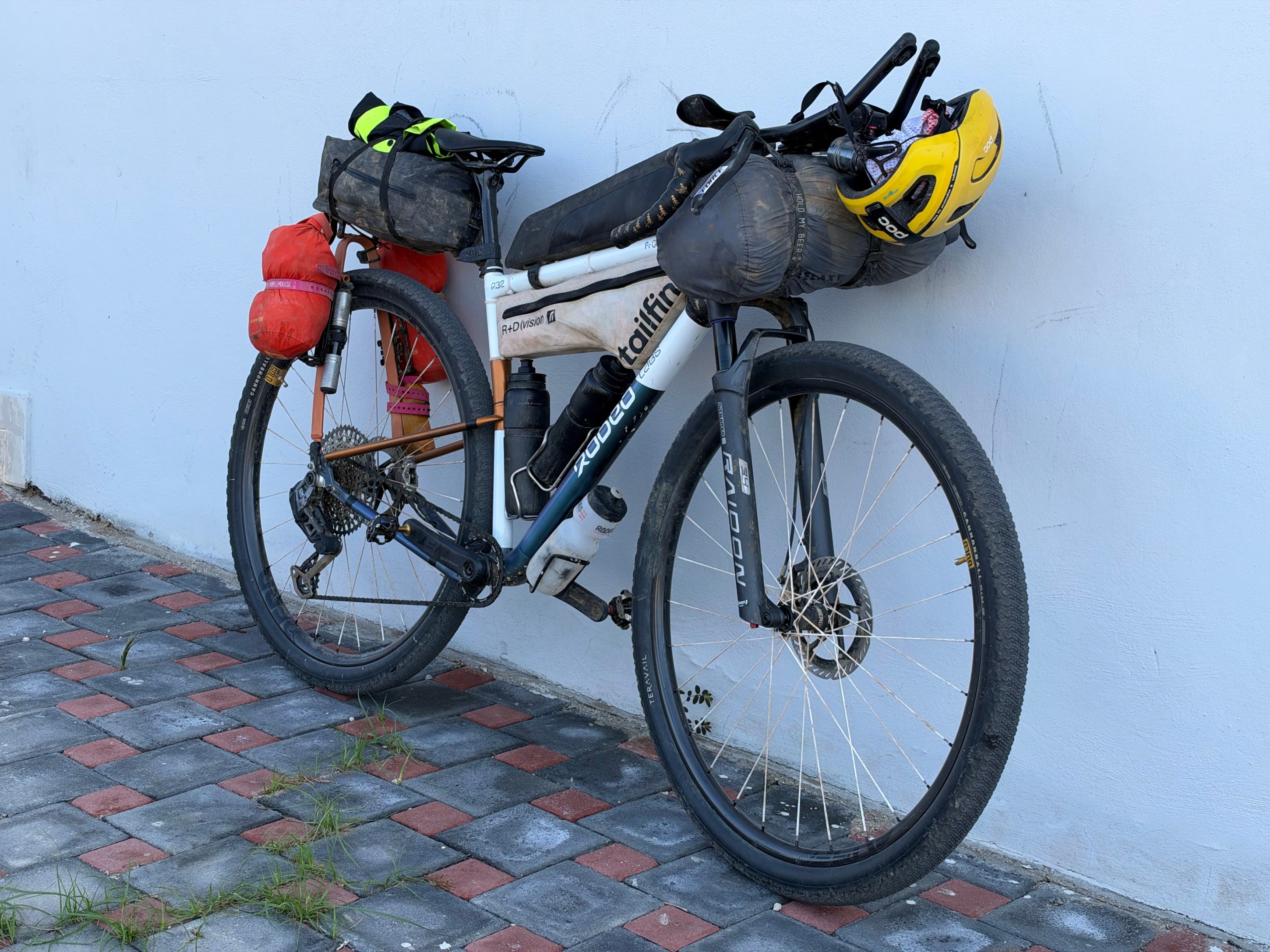 A bicycle equipped for touring, featuring bags attached to the frame, a helmet resting on top, and parked against a light-colored wall.