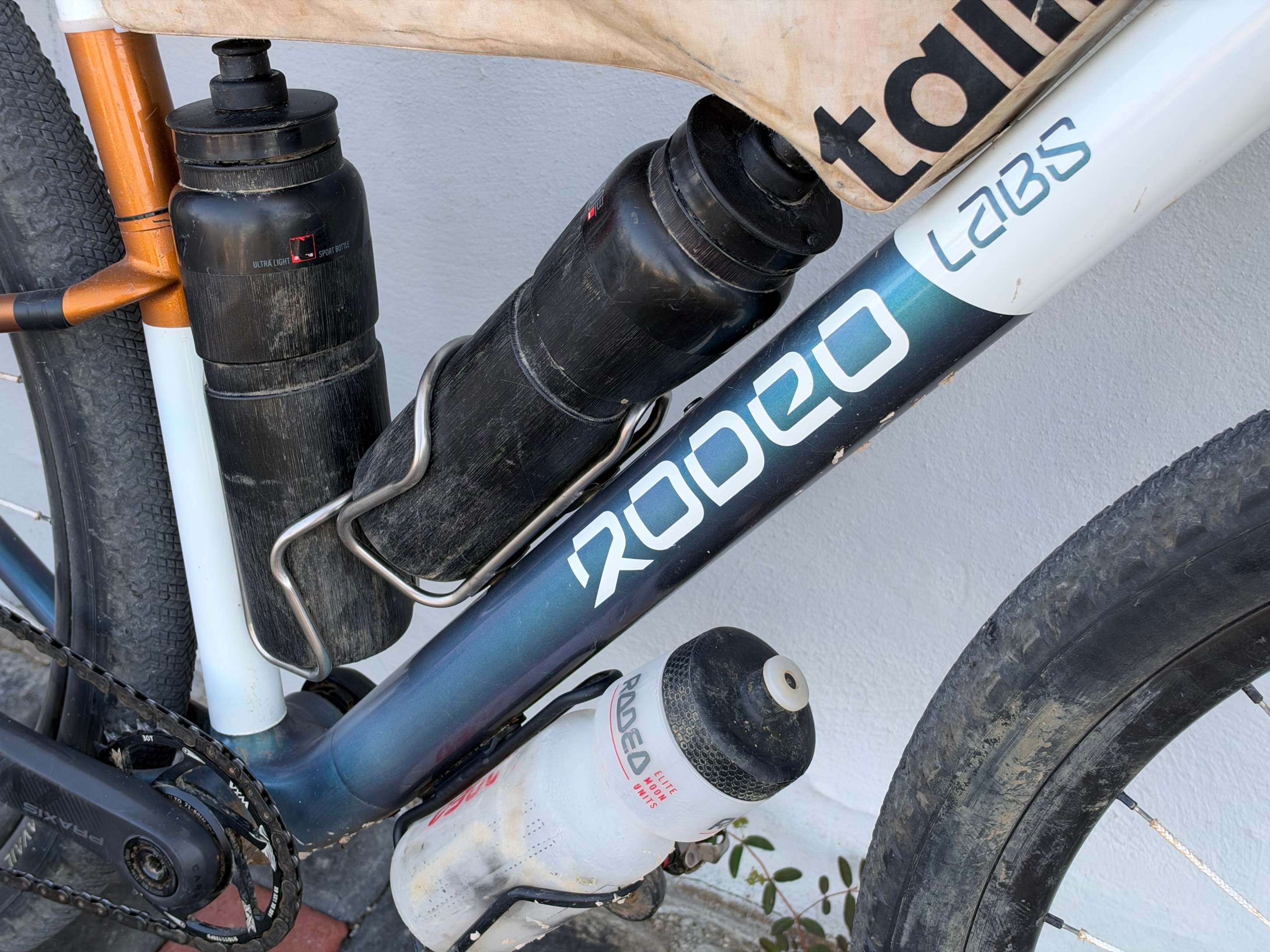 Close-up of a bicycle frame showcasing two water bottles attached, with one vibrant blue frame labeled 'Rodeo Labs' and visible dirt on the components.