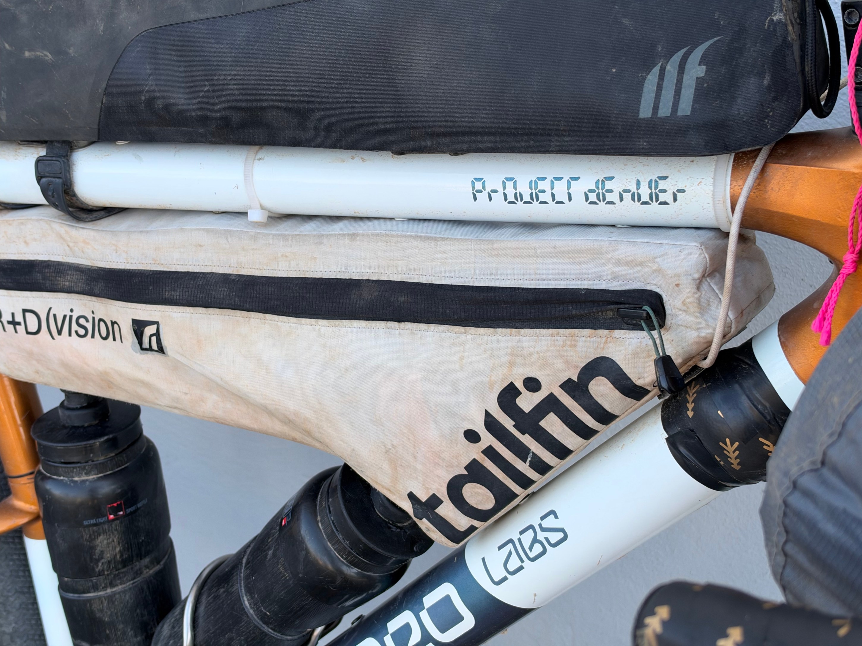 Close-up of a bicycle frame with various bags attached, including a Tailfin bag and a protective cover. Some components are slightly dirty, indicating usage.