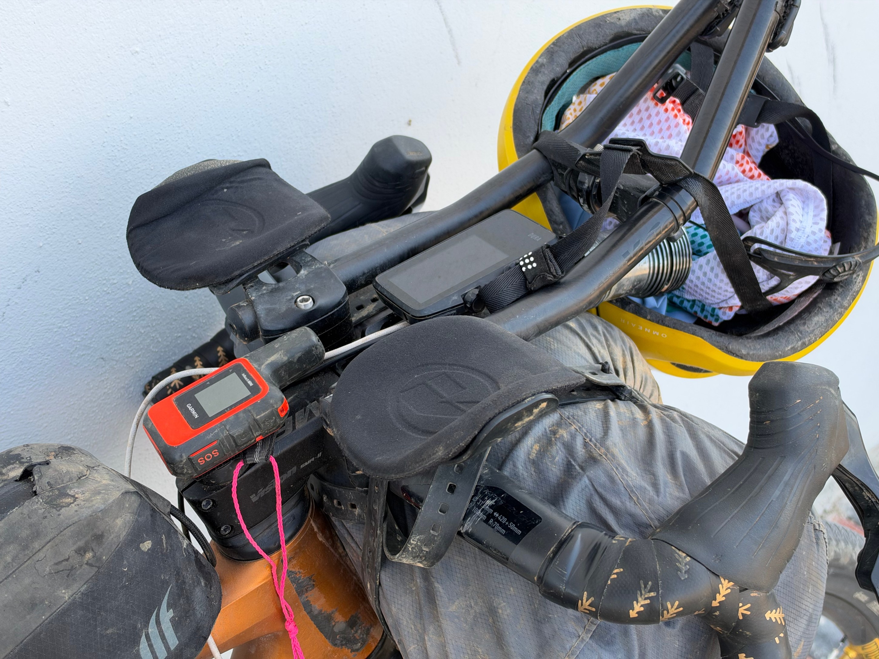 Close-up of a bicycle handlebar featuring a GPS device, gloves, and a helmet with gear straps, set against a light background.
