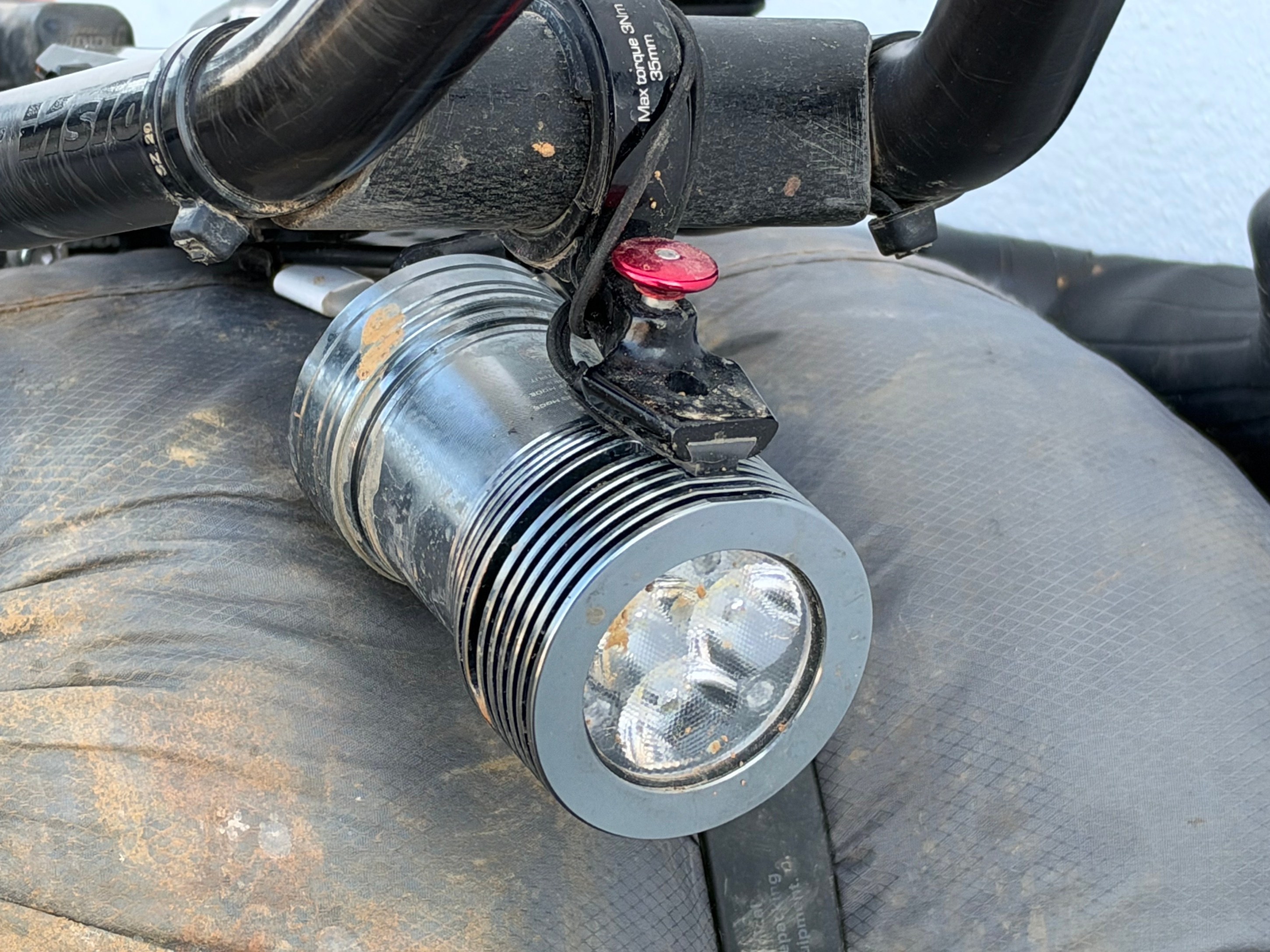 Close-up of a bicycle handlebar featuring a mounted LED flashlight, partially covered in dirt.
