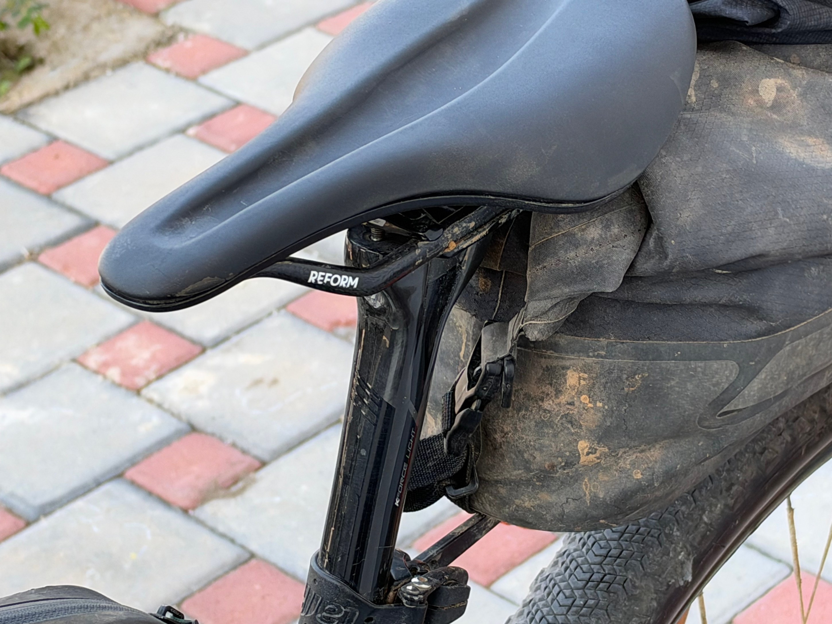Close-up of a bicycle saddle labeled 'REFORM' attached to a bike, with a dirty seat bag and textured tire visible in the background.